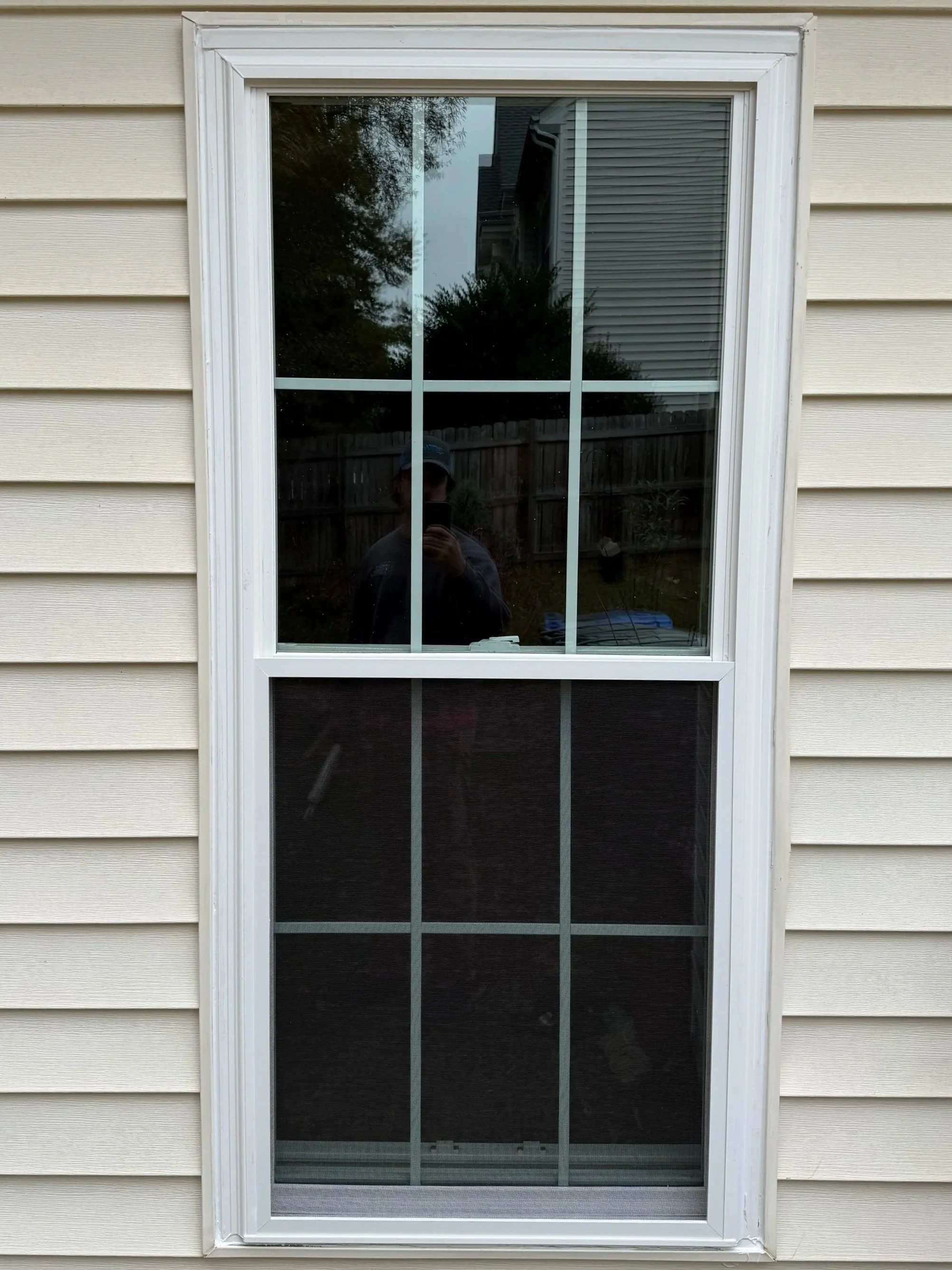 A window with decorative grid panes on cream-colored siding, with a person taking a photo reflected in the glass.