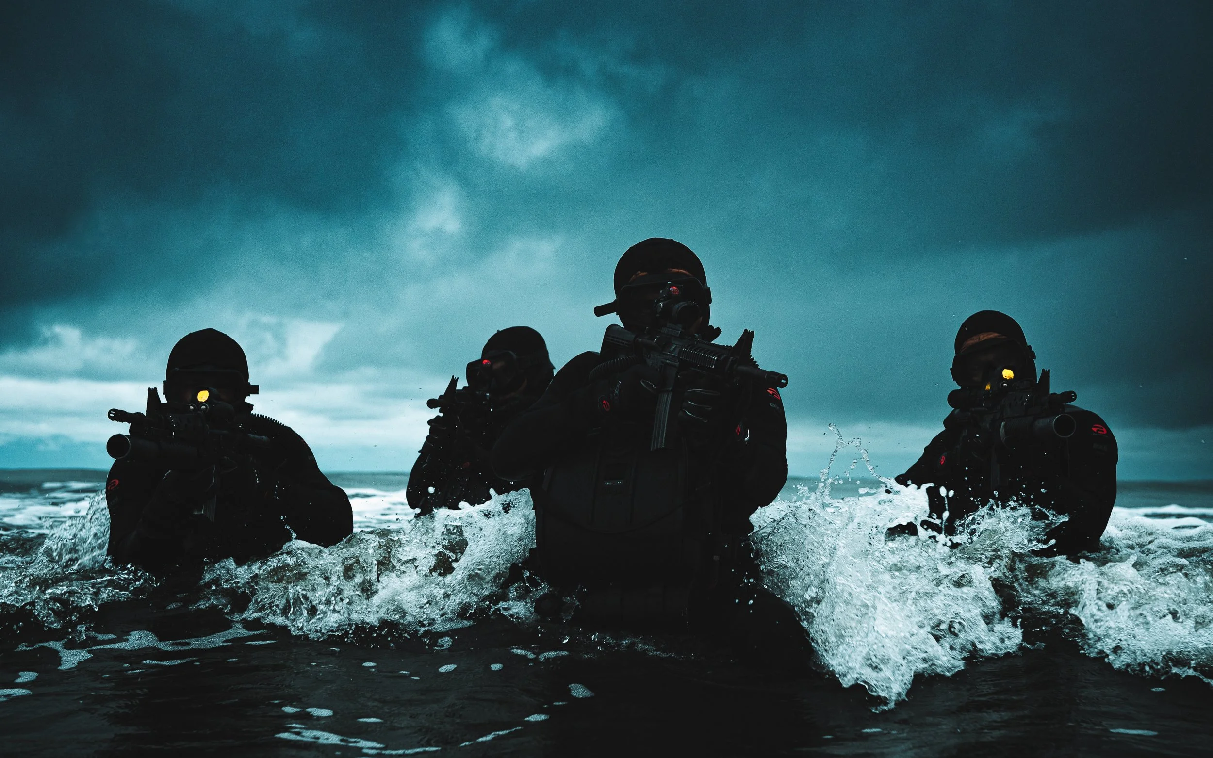 Four soldiers in tactical gear wading through rough surf under dark clouds