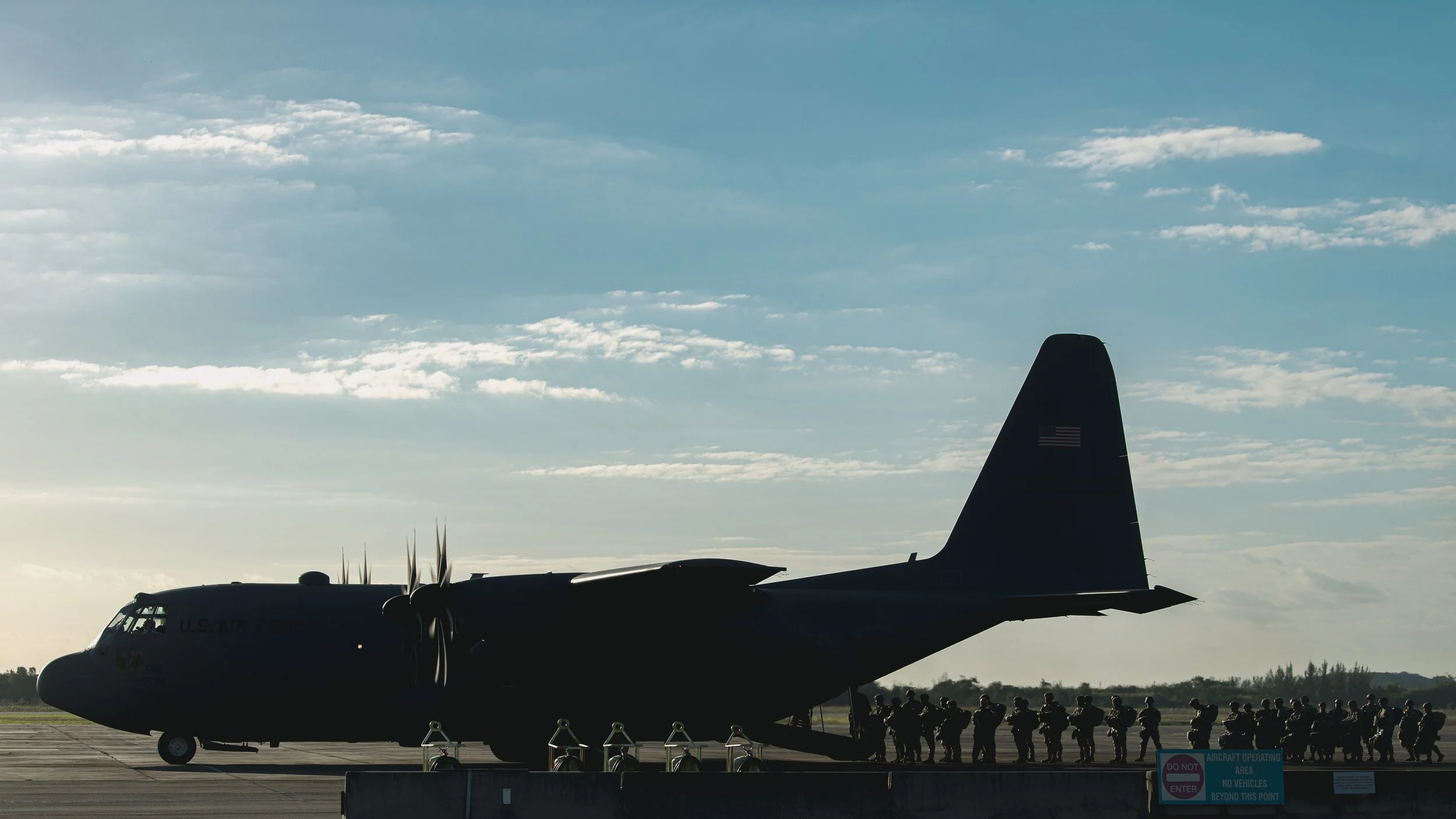 Silhouette of a military transport aircraft on the tarmac, troops lined up beside it under a cloudy sky