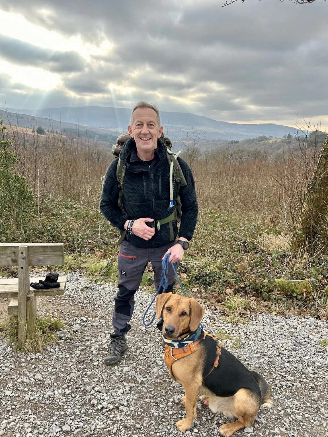 A smiling man in outdoor hiking gear stands with a dog on a trail in a hilly, wooded area, with mountains and a cloudy sky in the background.