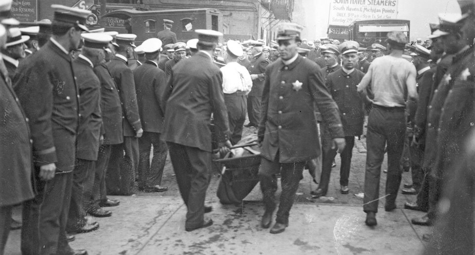 Crowd of police officers and civilians gathered in a city street, with some officers pushing a cart or stretcher, likely from an early 20th-century event.