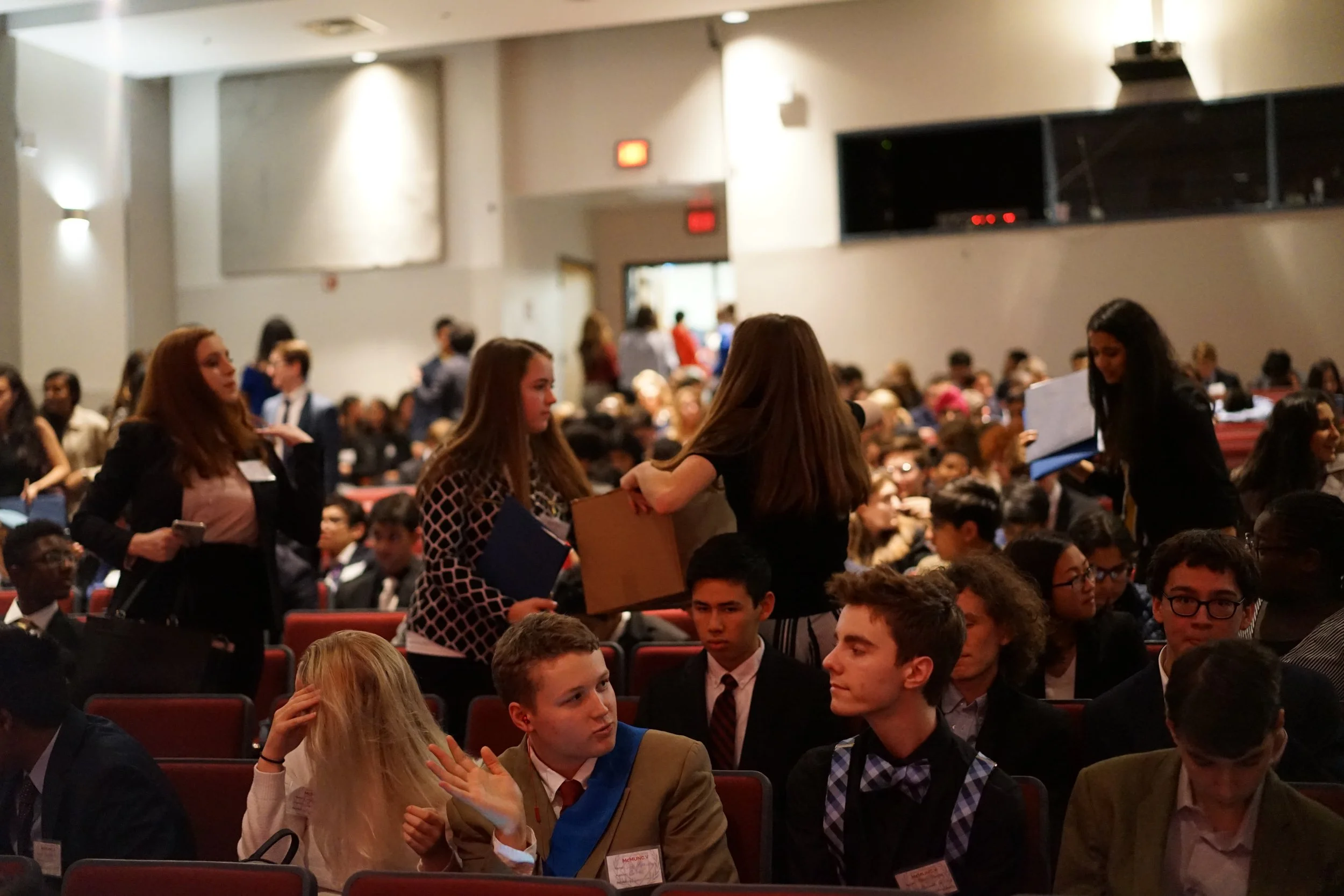 A large group of young people gathered in an auditorium or conference hall, some standing and talking, others seated, with a stage or presentation area in the background under dim lighting.