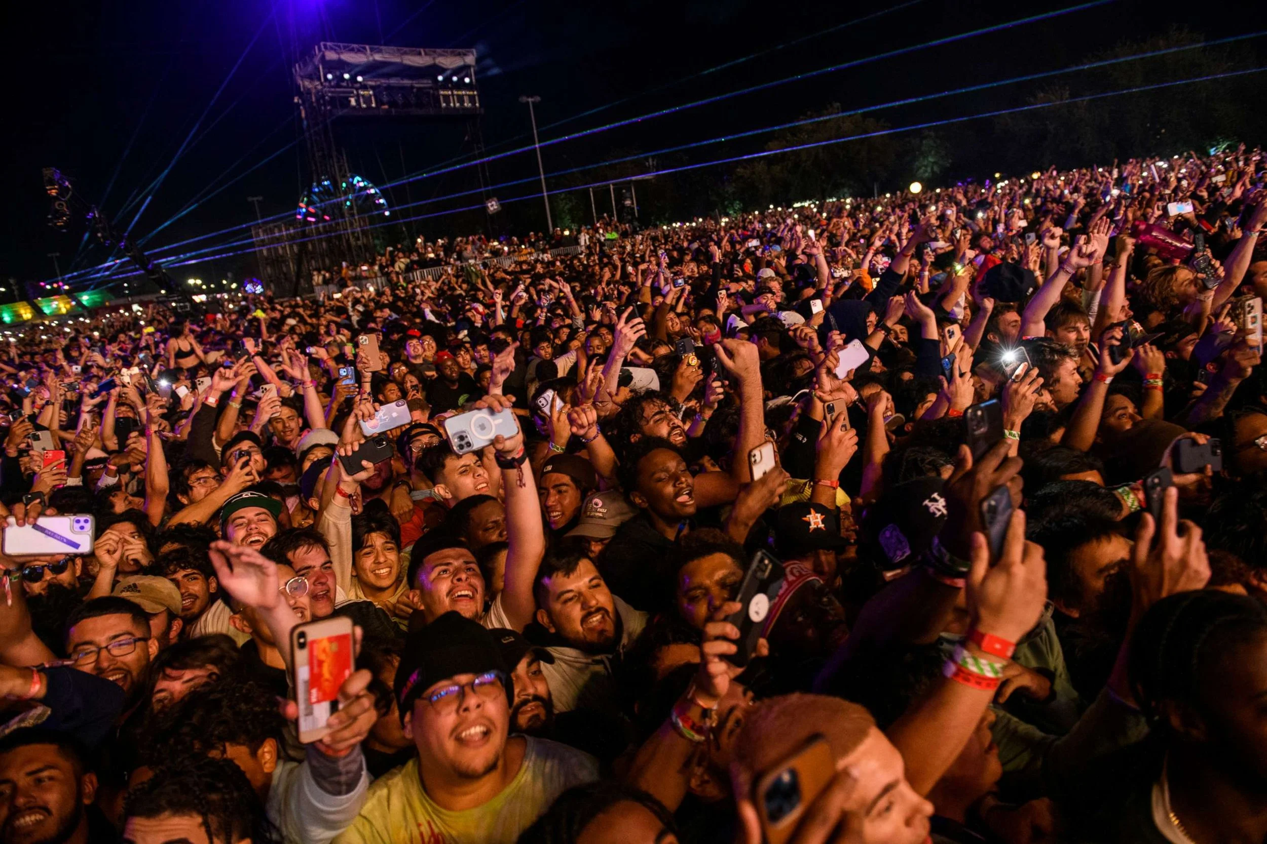 A large crowd of people at a nighttime concert or festival, many holding up phones and taking pictures, with laser lights and stage lighting in the background.