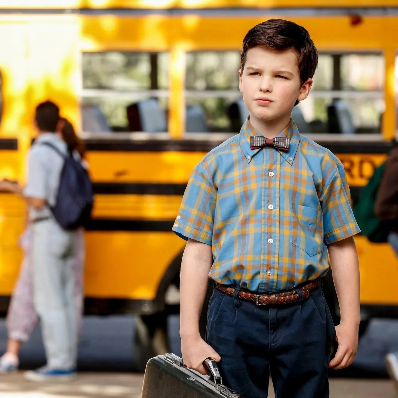 A young boy with dark hair, wearing a plaid yellow and blue shirt and a bow tie, stands in front of a yellow school bus, holding a briefcase.