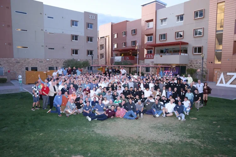Large group of people gathered outdoors on a grassy area in front of apartment buildings, posing for a group photo during daytime.