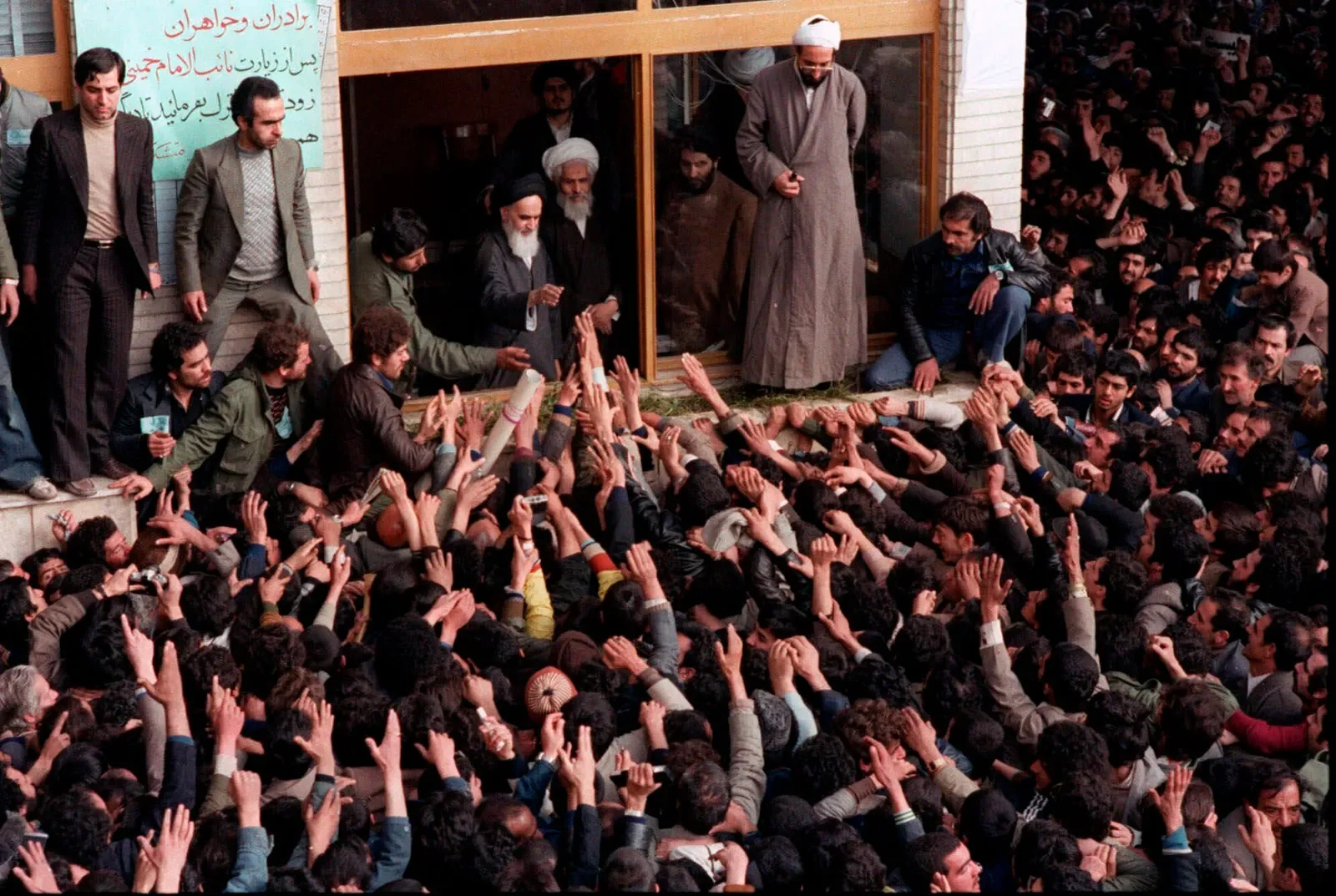 Large crowd of people gathered in front of a building, many reaching out their hands contact with others, possibly in a demonstration or rally. Several men are standing on the building's steps and also on the ground, some overseeing the crowd.