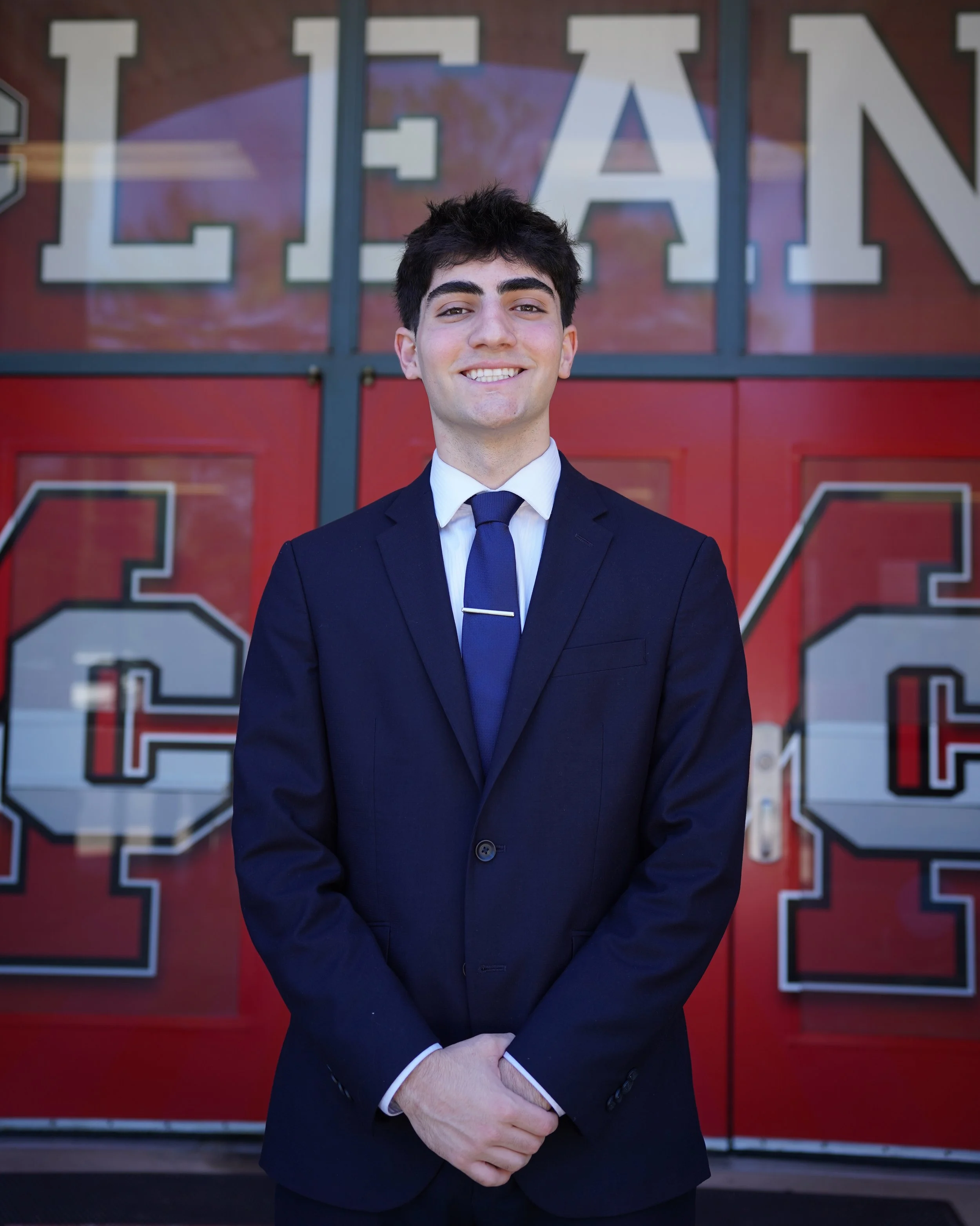 A young man in a formal suit with a tie, standing in front of a red background with large white letters. He is smiling and has dark, slightly messy hair.