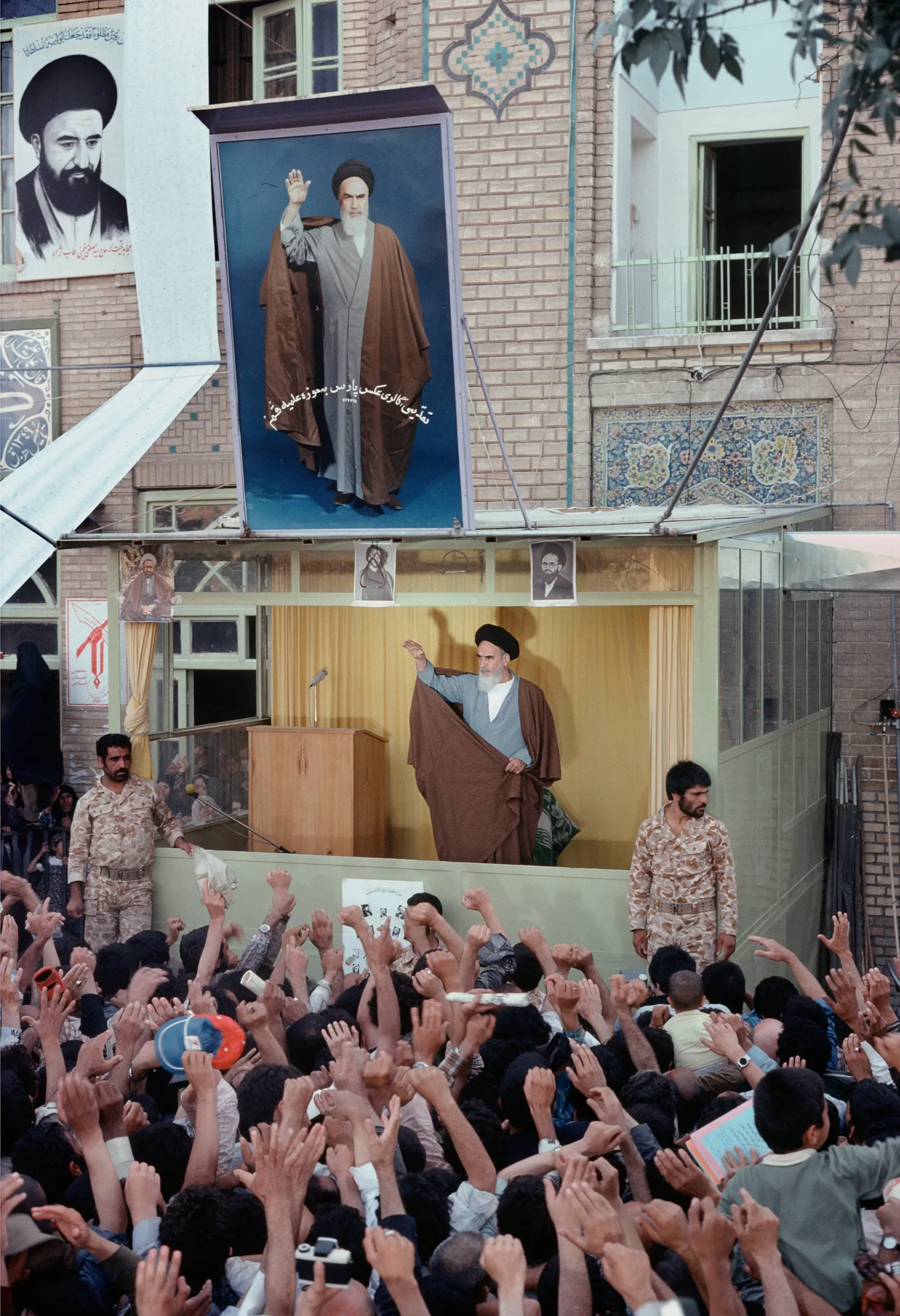 Cleric on stage speaking to a large crowd, with protesters raising hands and taking photos, buildings in the background, banners and portraits displayed.