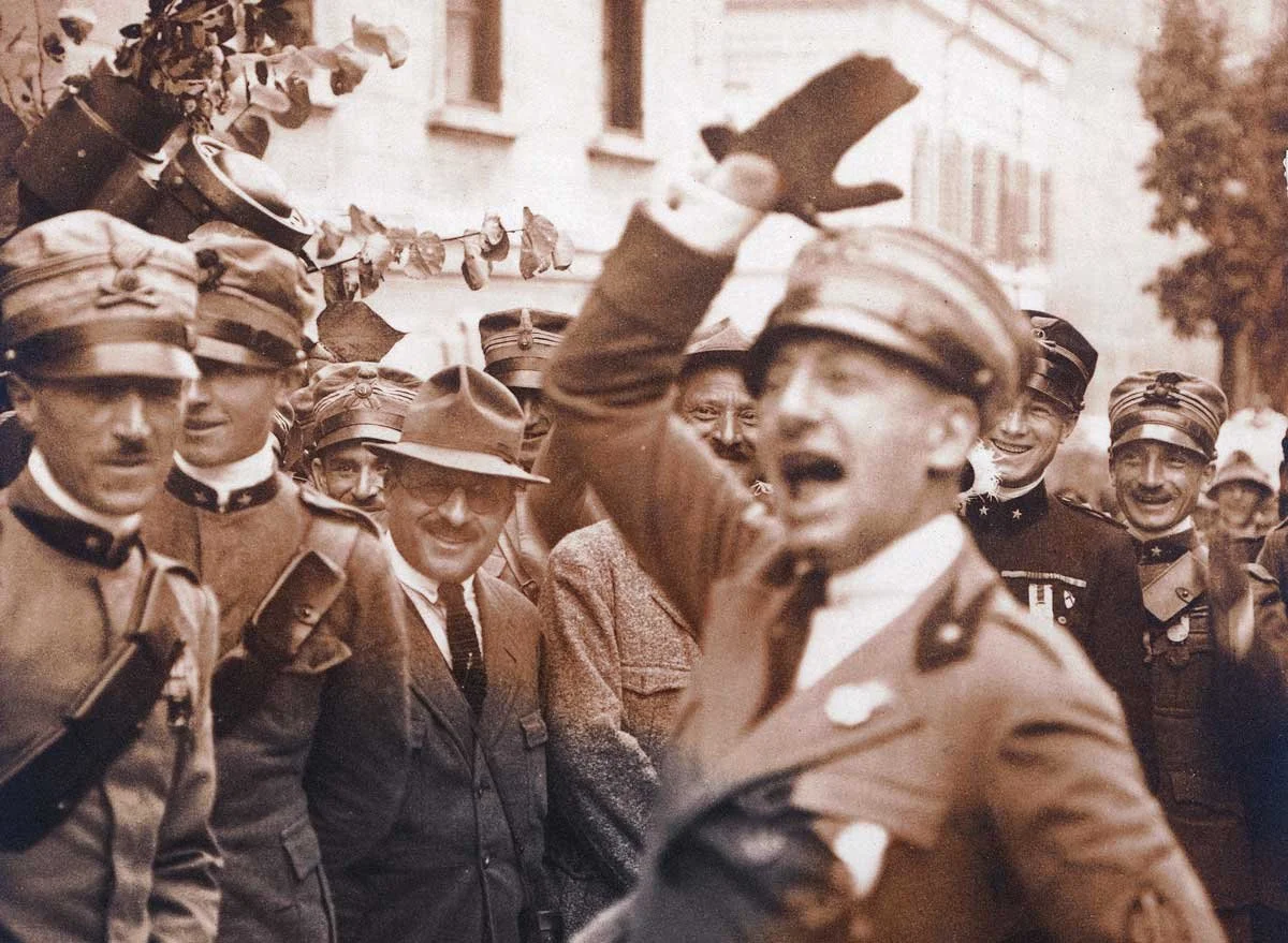A black and white photo of a group of military officers and civilians, some smiling and some laughing, with one man in the foreground saluting or waving.