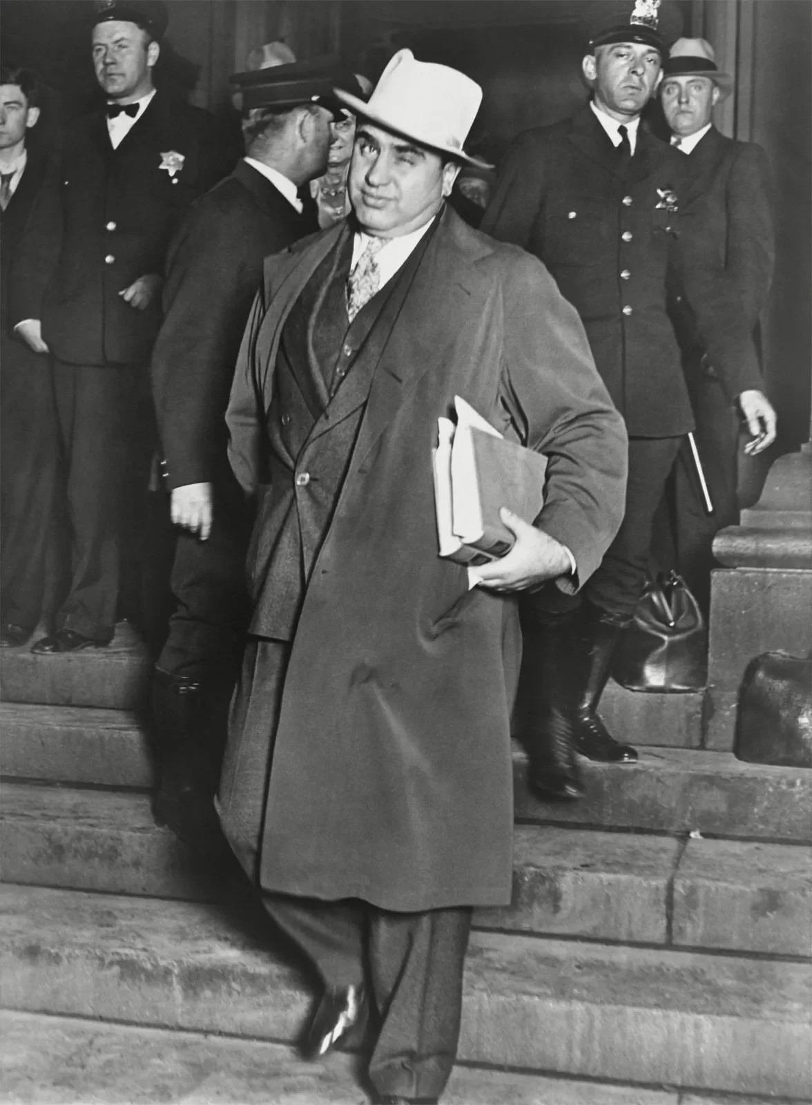 Black and white photo of a man in a suit and trench coat with a white hat holding folders, walking down stairs with a confident expression. Several men in suits and police uniforms are in the background.