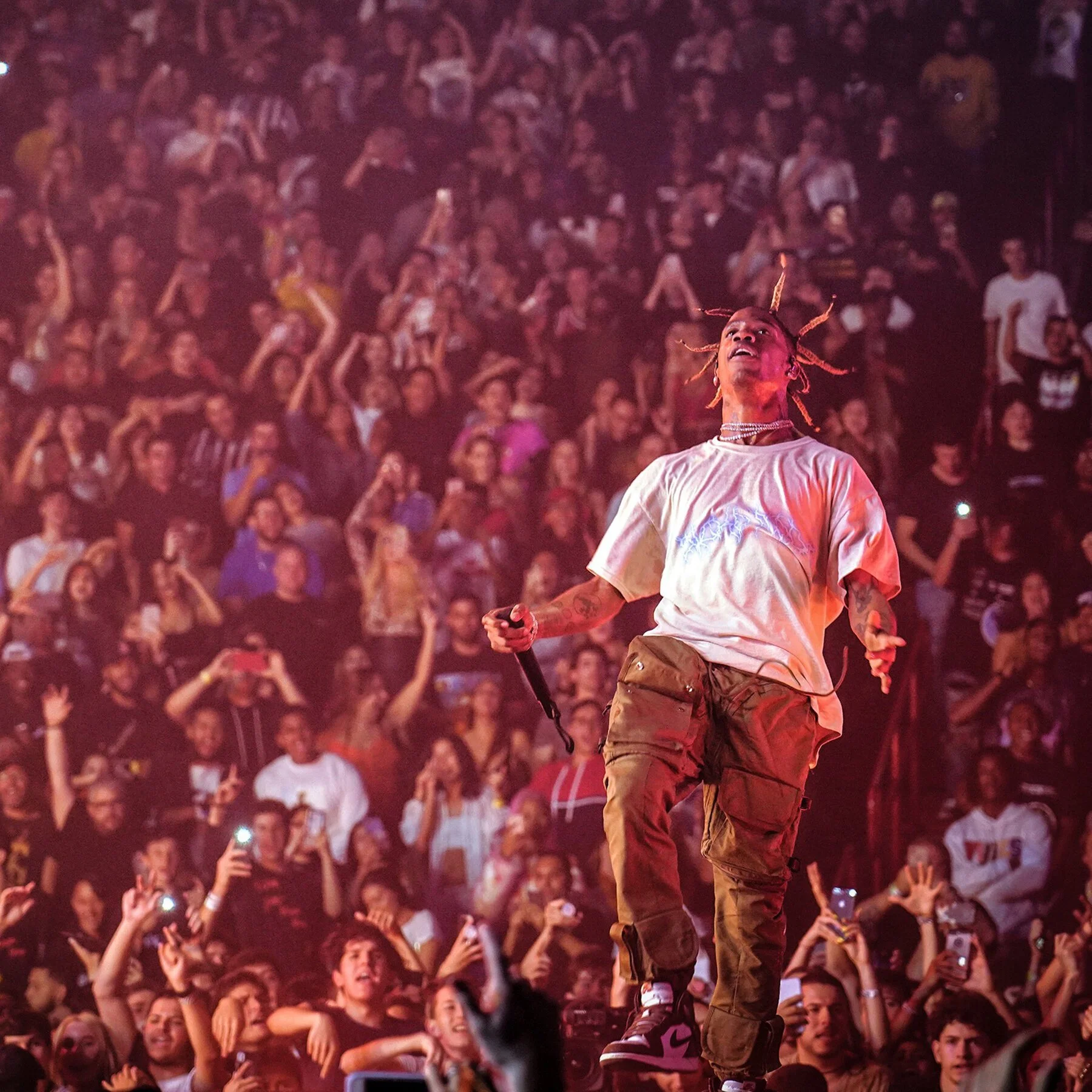 A performer with braided hair and tattoos on his arm stands on stage with a microphone, surrounded by a large audience at a concert or music event.