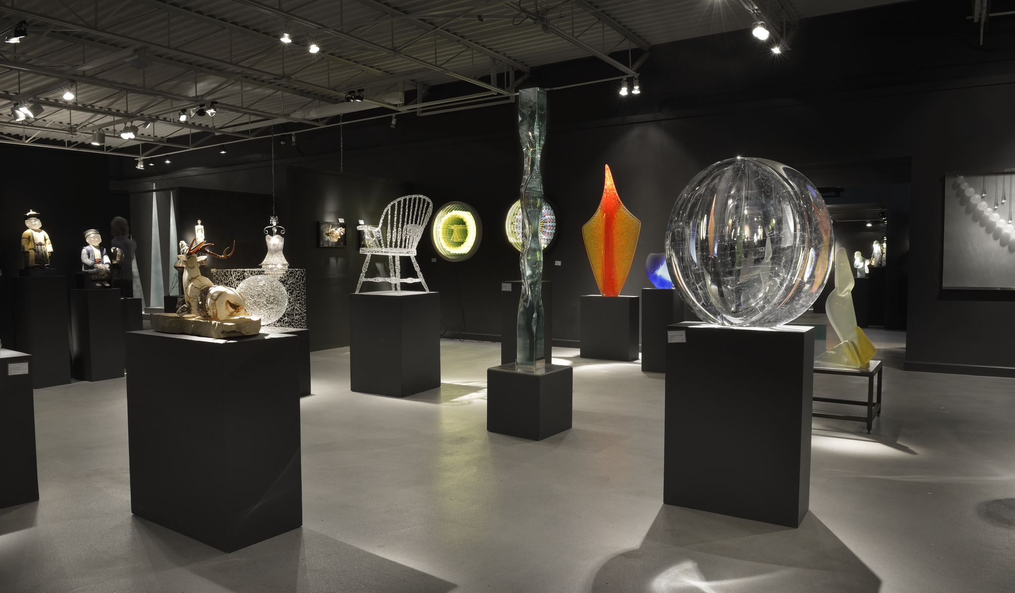Art gallery exhibit featuring various glass sculptures and figurines displayed on black pedestals in a dimly lit room.