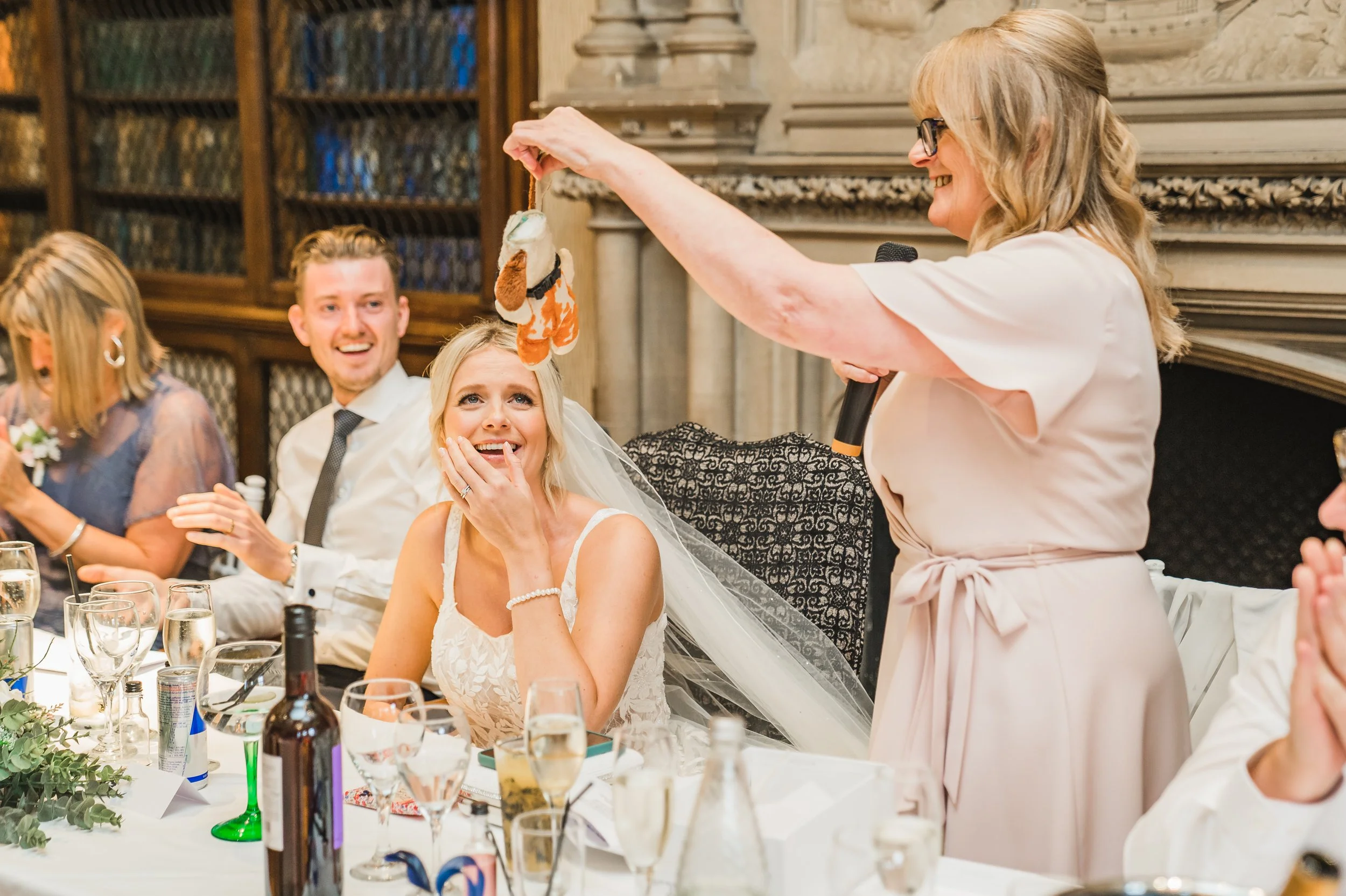 A smiling bride during a wedding reception. The bride, seated, is touching her face and looking up at the toy with amusement. Other guests are visible at the table, which is decorated with drinks and greenery, at The Elvetham.