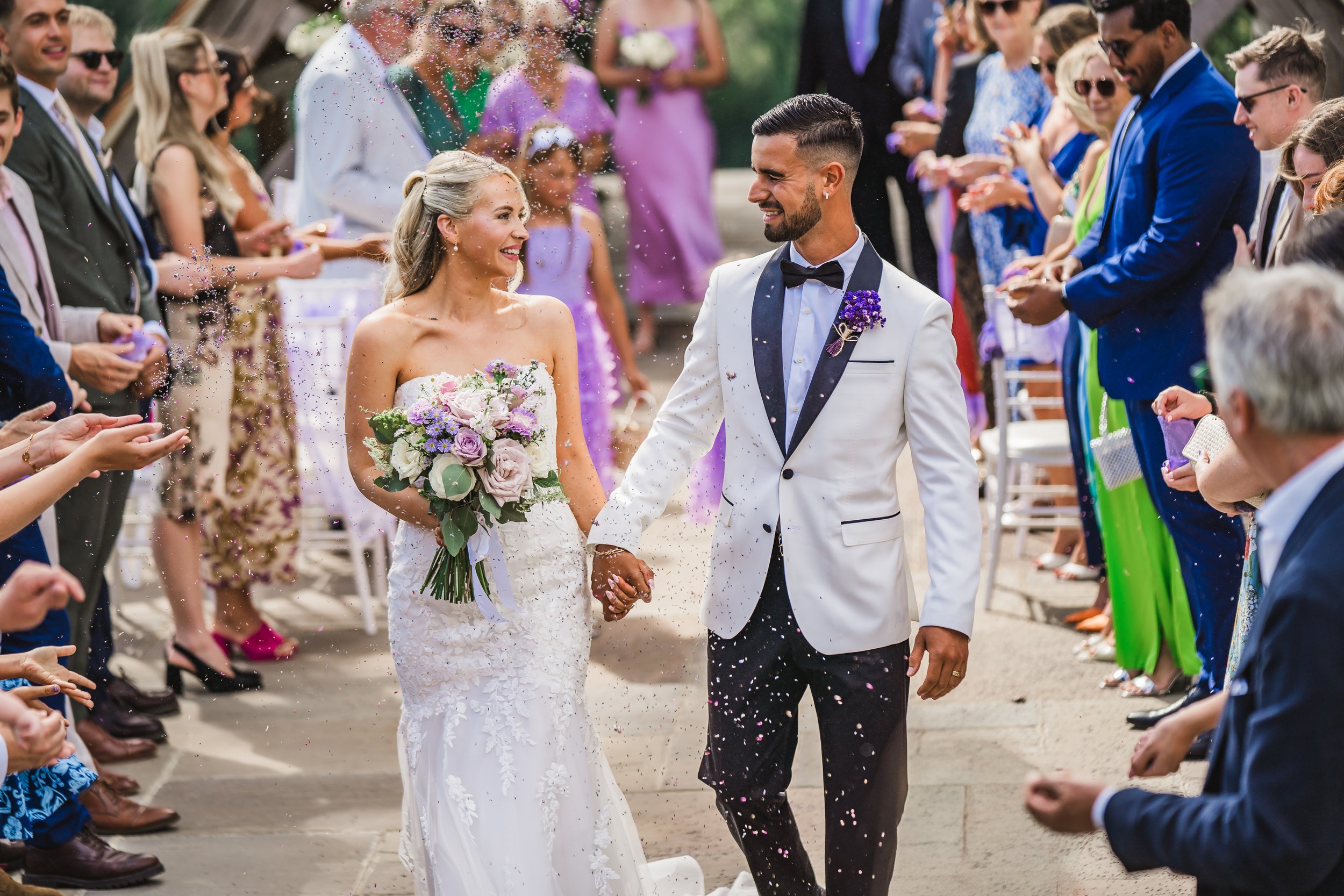 A bride and groom holding hands walking through a crowd of wedding guests outdoors during the wedding celebration.