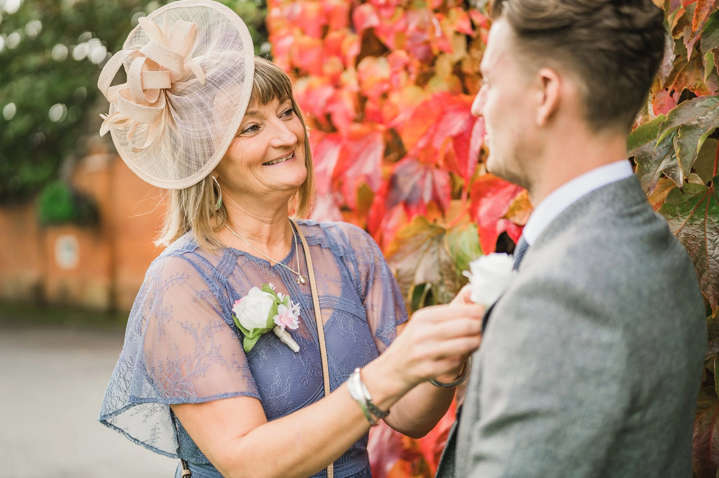 A woman wearing a large, beige, decorative hat and a purple lace dress is smiling at a man dressed in a gray suit. She is holding a small bouquet of flowers, and they are standing outdoors in front of a colorful backdrop of red and orange leaves, sug