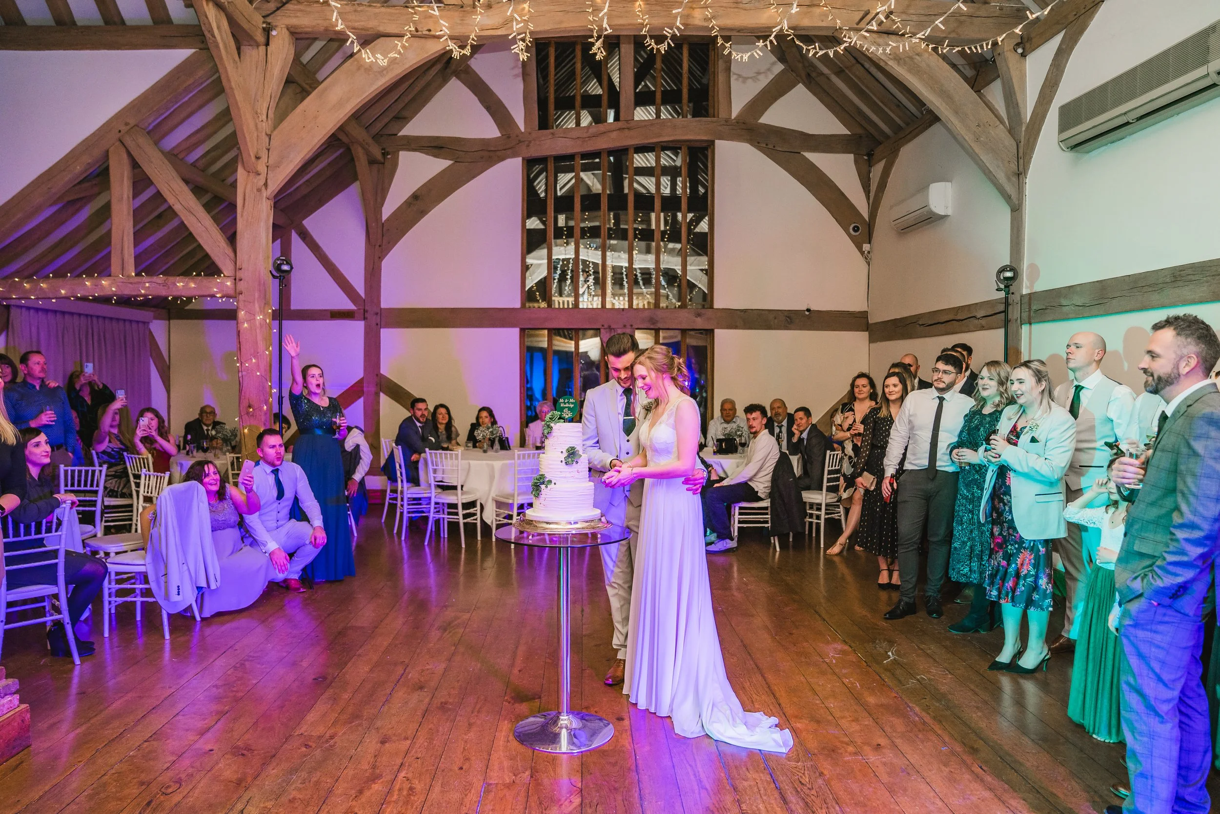 A wedding reception inside a rustic barn with exposed wooden beams and string lights. The bride and groom are cutting a tiered white wedding cake at a high table, surrounded by family and friends. Guests are seated at round tables and standing, watch