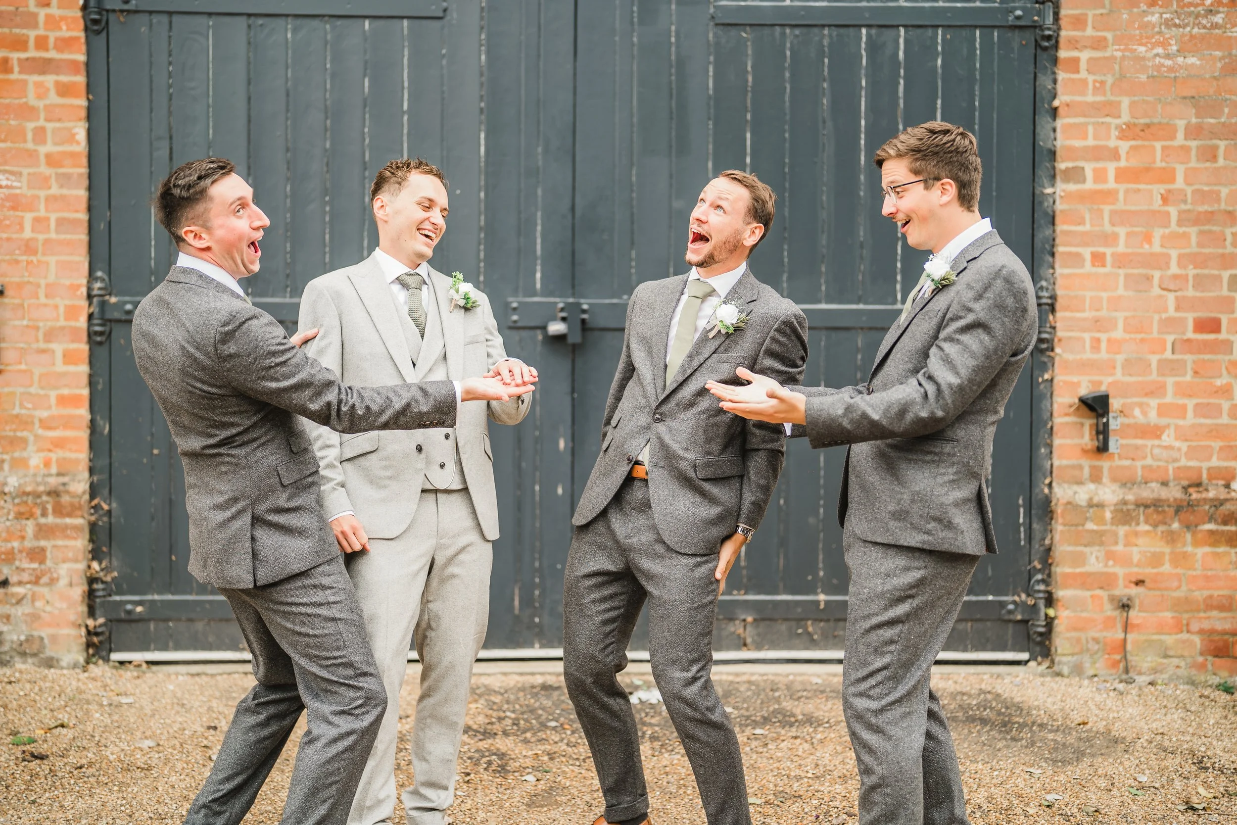 Four men dressed in suits laughing and chatting outside in front of a dark gray door with brick walls on either side.