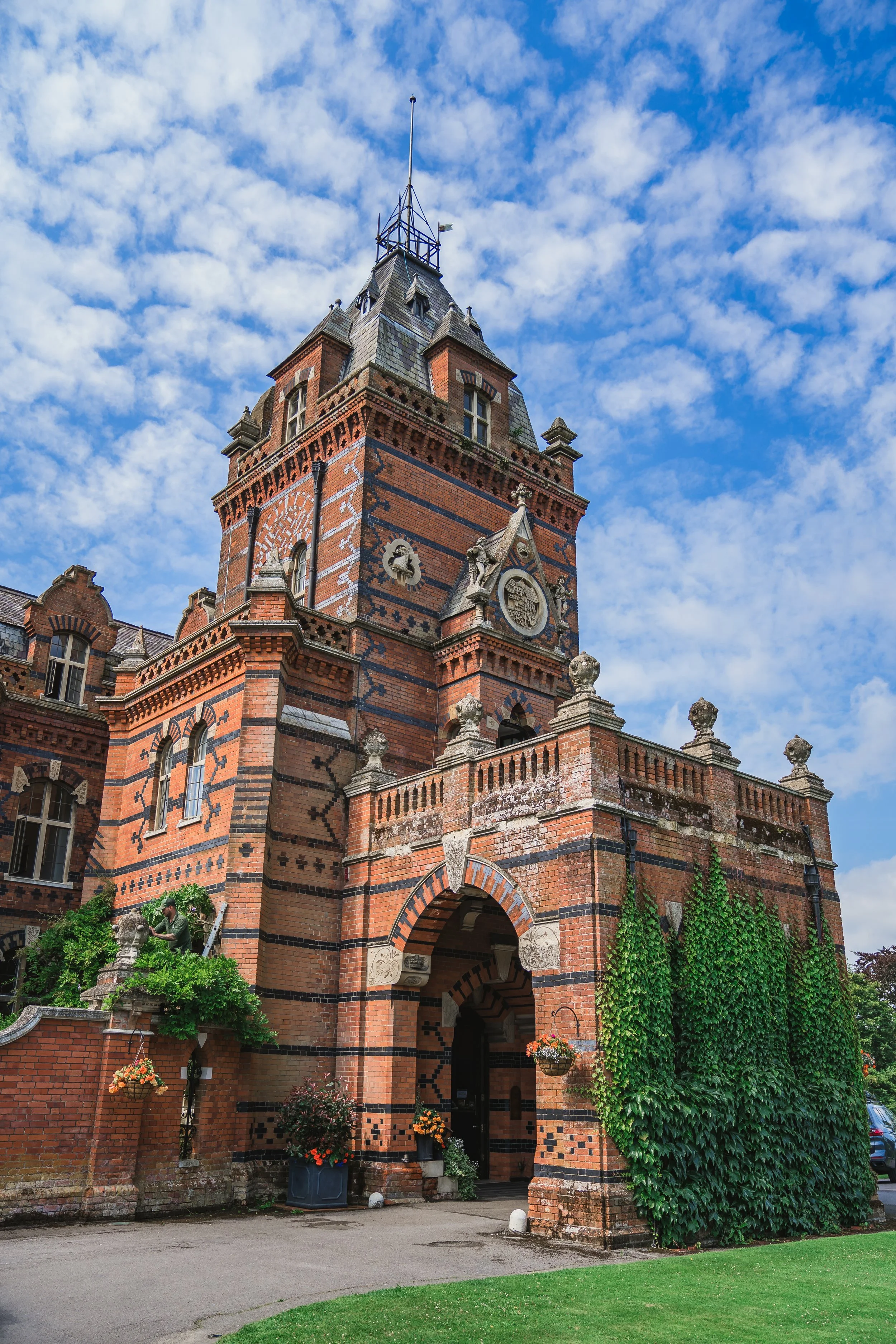 A historic red brick building with Gothic architectural features, ivy growing on one side, hanging flower baskets near the entrance, a man working on plants, and a blue sky with scattered clouds.