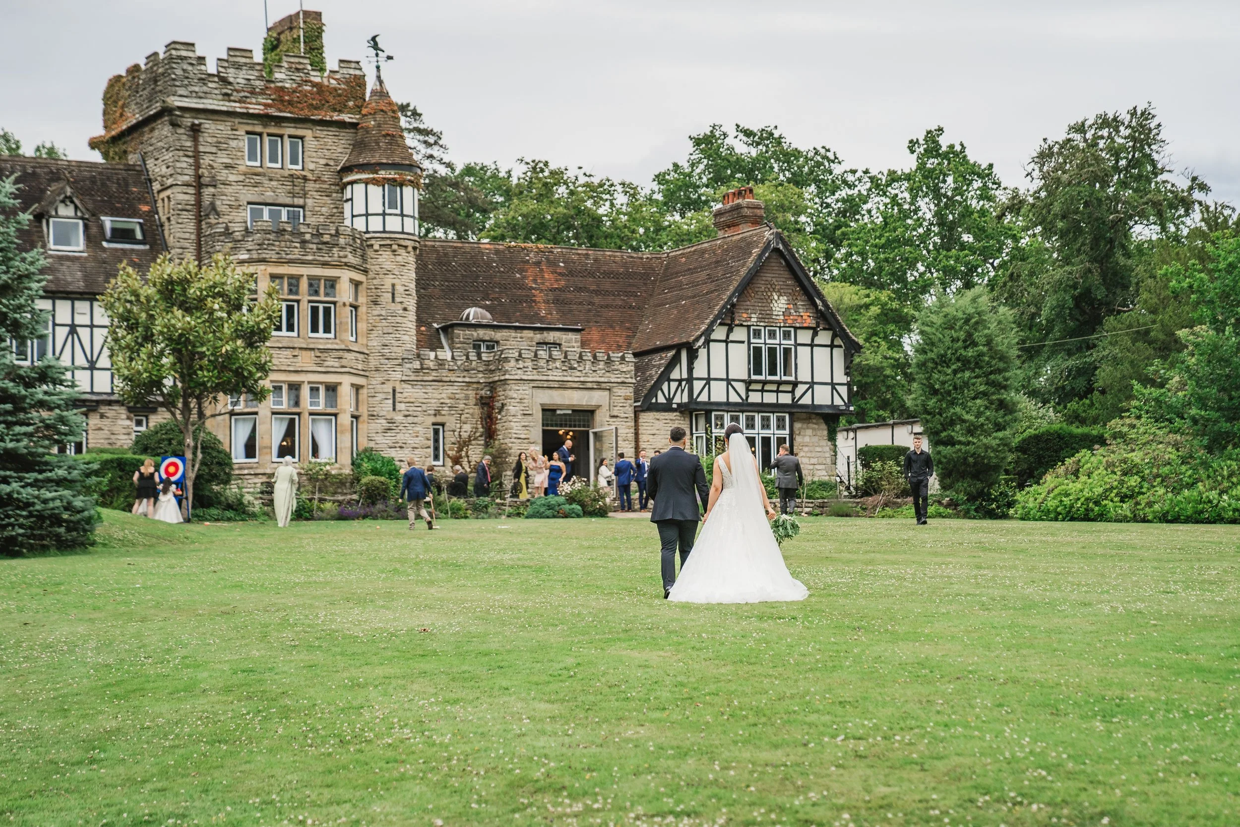 A bride and groom walk toward a group of wedding guests outside a large historic stone house with Tudor-style architecture, surrounded by lush green trees and a lawn. The Ravenswood