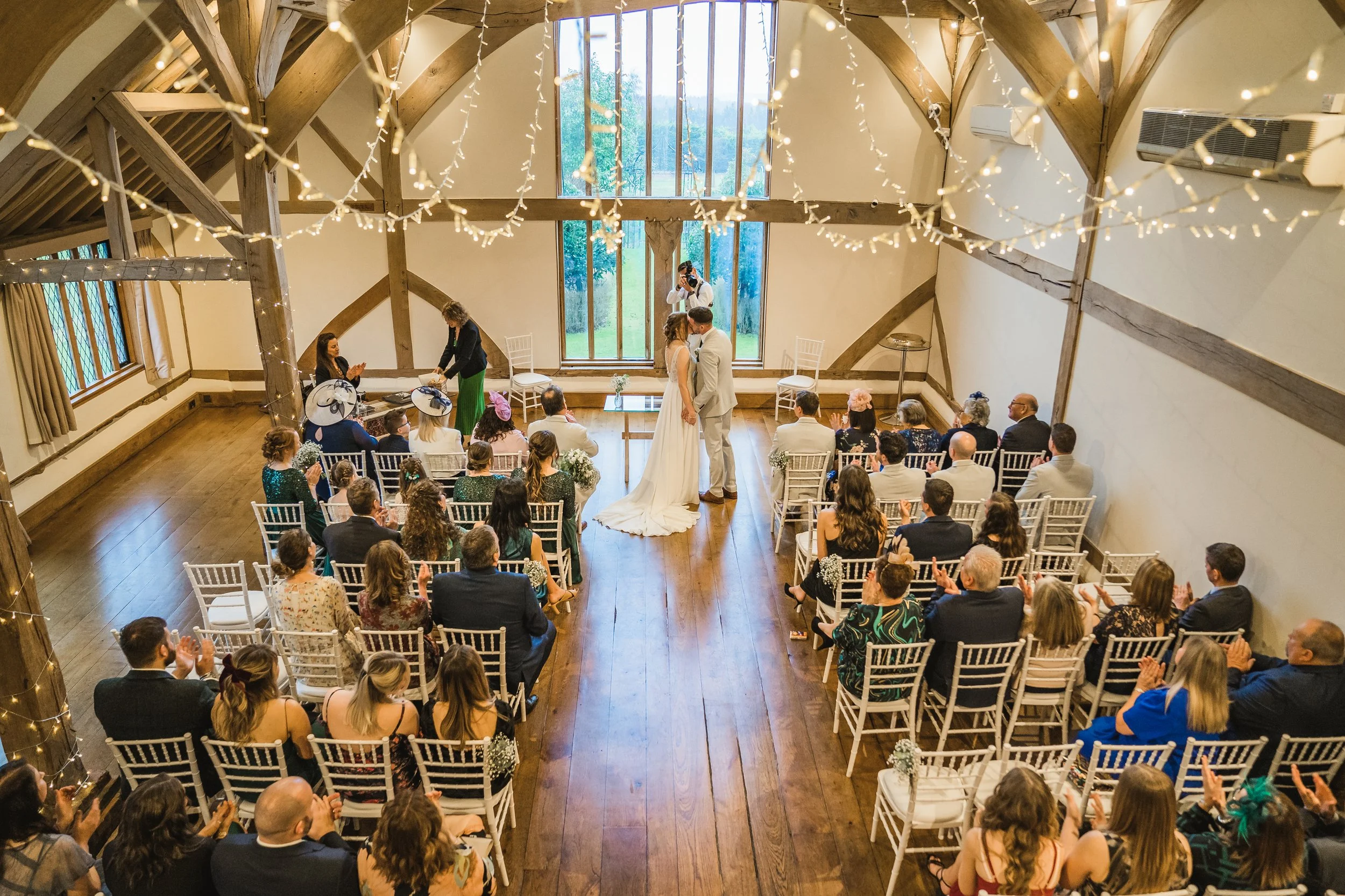 A wedding ceremony inside a charming wooden barn with guests seated and the bride and groom kissing. Decor includes string lights and large windows with a view of greenery outside.