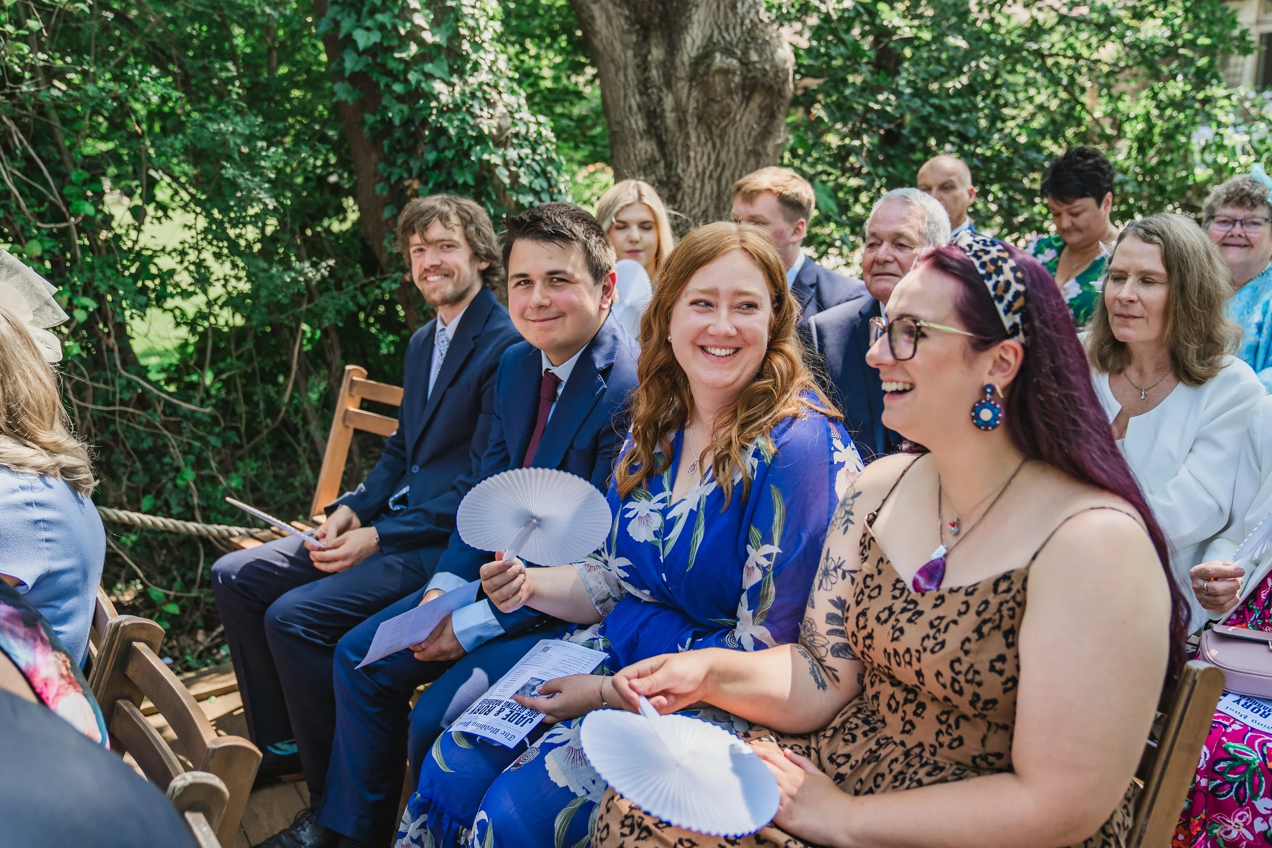 People seated outdoors in chairs, attending an event or ceremony, smiling and holding fans, with green foliage and trees in the background.