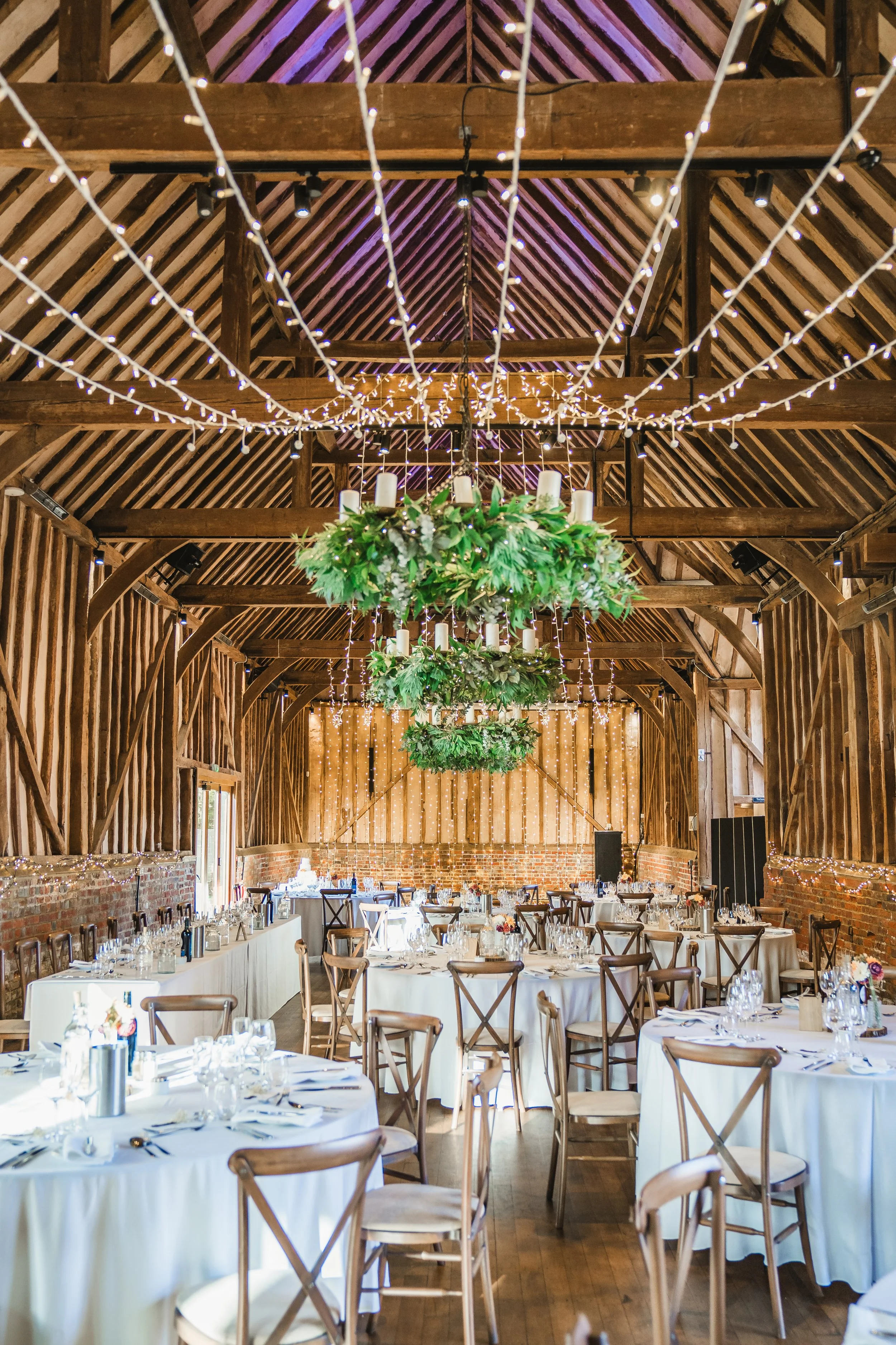 Event space decorated with hanging string lights, chandeliers with greenery, and round tables with white tablecloths set with glassware, silverware, and wine bottles, within a rustic wooden barn with exposed beams and brick accents.