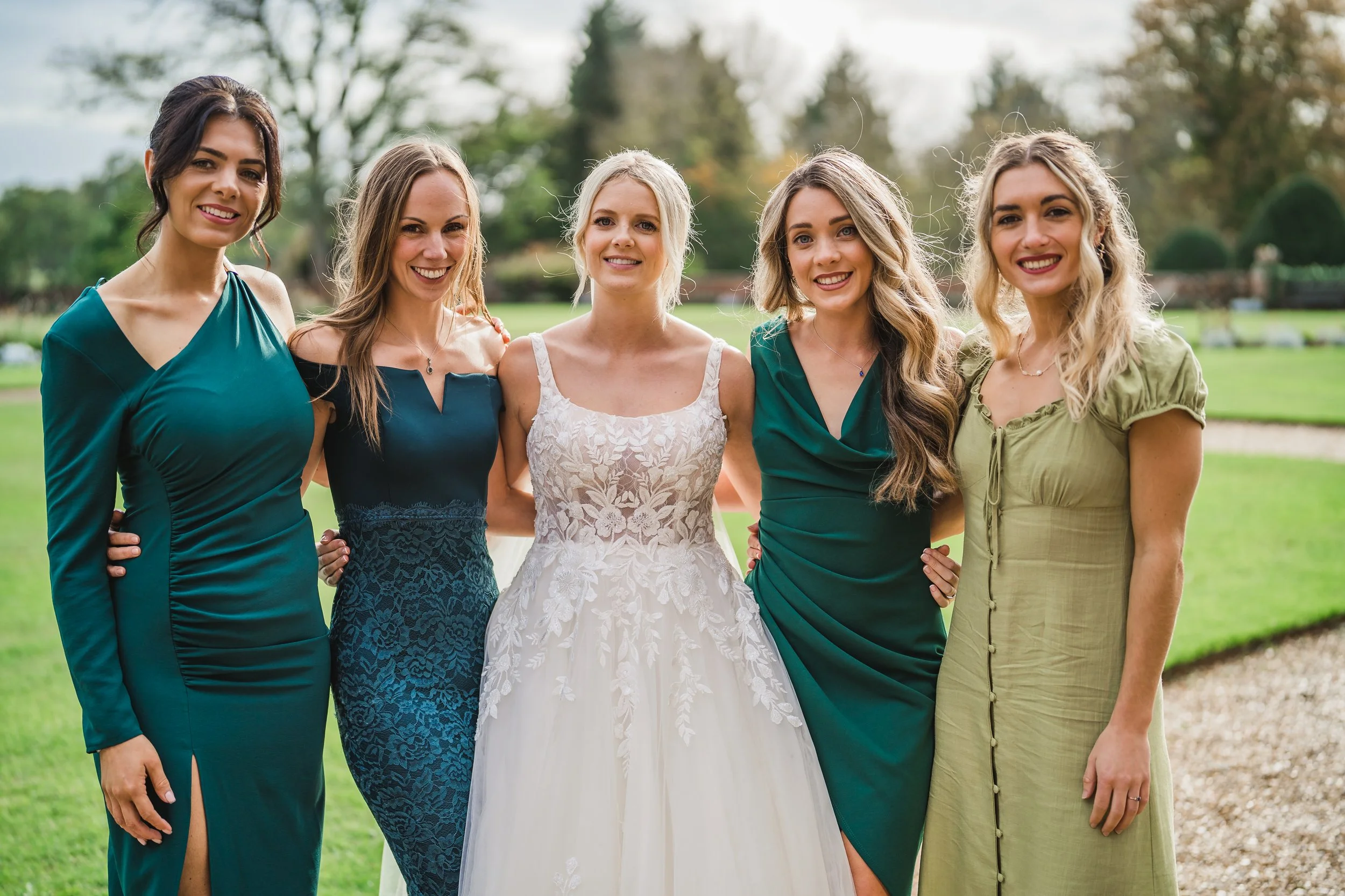 Group of six women smiling outdoors, one in a white lace wedding dress, with trees and grass in the background.