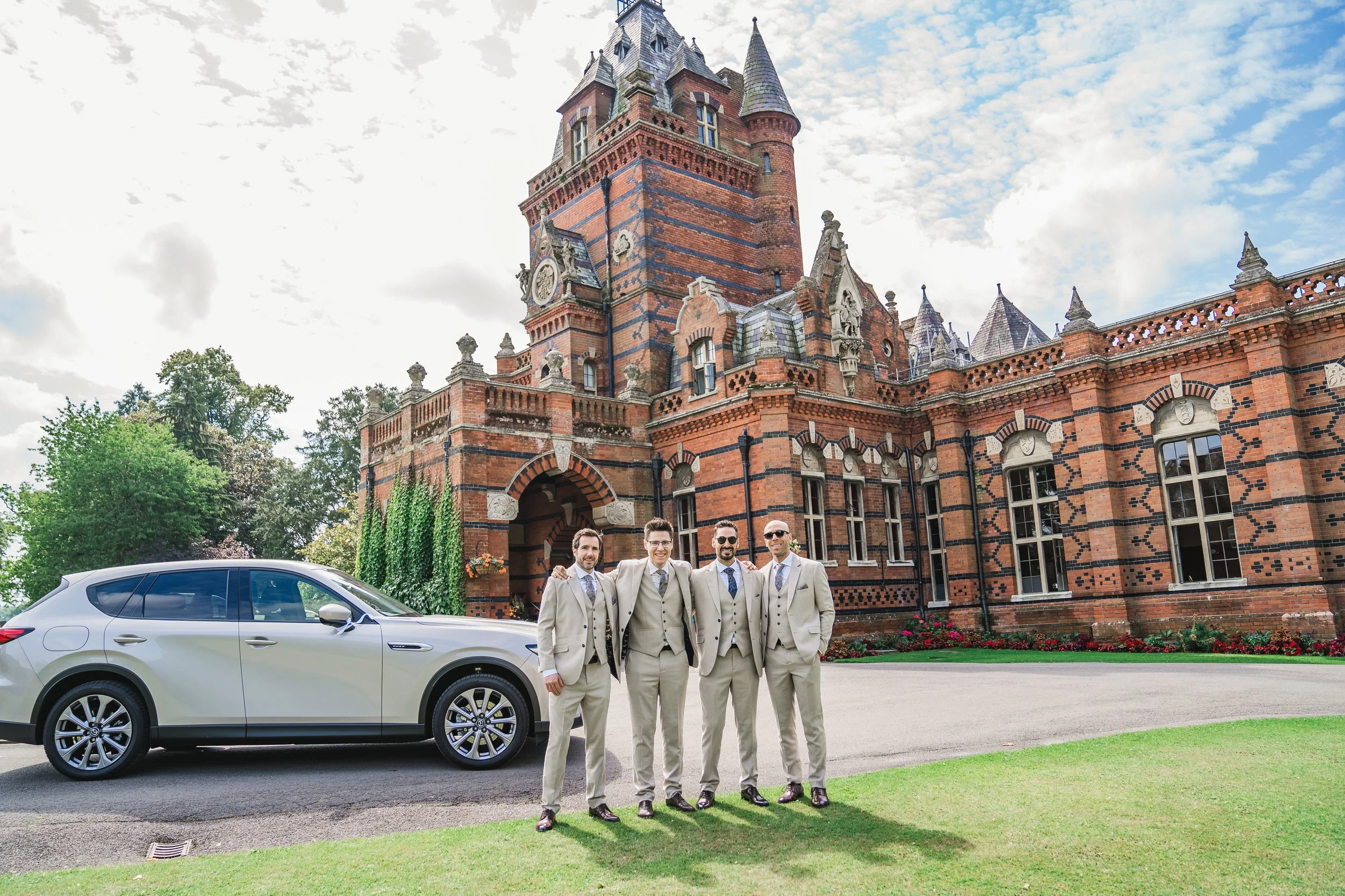 A group of five men dressed in beige suits standing arm-in-arm in front of a large red brick castle-like building, with a modern silver car parked nearby and green trees surrounding the scene.