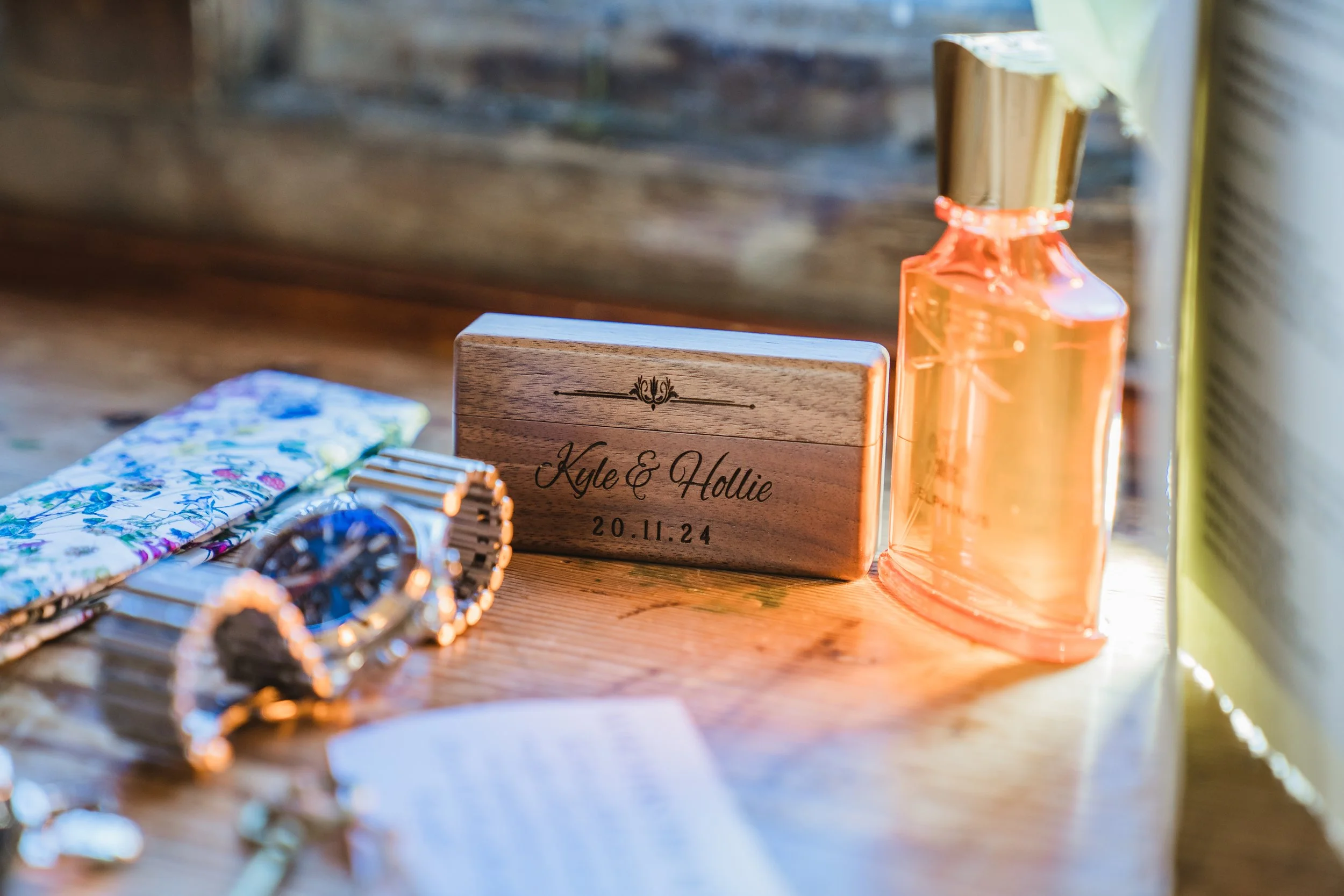 A wooden table with a wooden block engraved with the names 'Kyle & Hollie' and the date '20.11.24,' gold-colored bottles, a floral-patterned cloth, and a silver watch with a chain, illuminated by natural light.