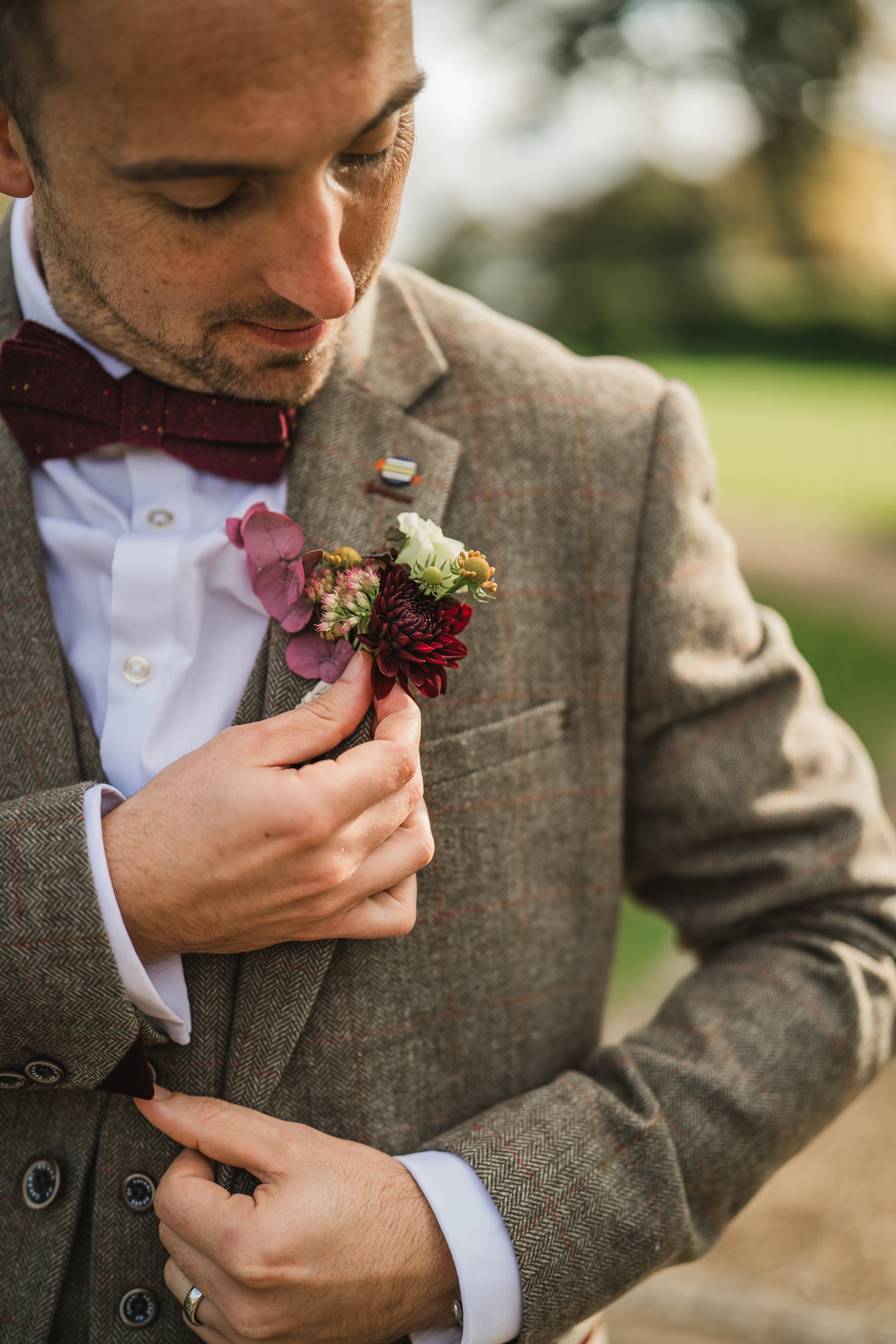 A man in a brown plaid blazer, white shirt, and red bow tie is holding a small boutonniere with white, red, pink, and purple flowers close to his chest. The background is a blurred outdoor setting with trees and grass.