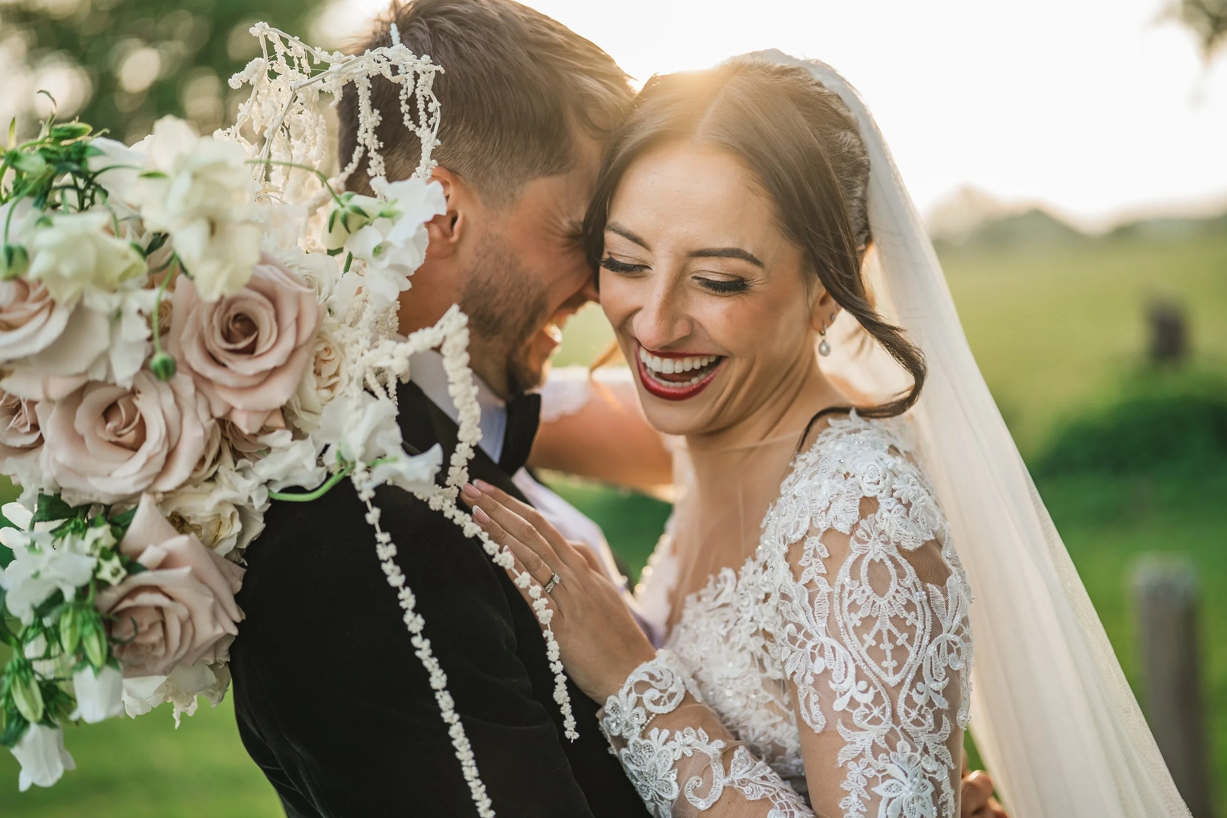 A bride and groom smiling while leaning their foreheads together outdoors during sunset, with the bride wearing a lace wedding gown and the groom in a black tuxedo, surrounded by greenery. Burley Manor