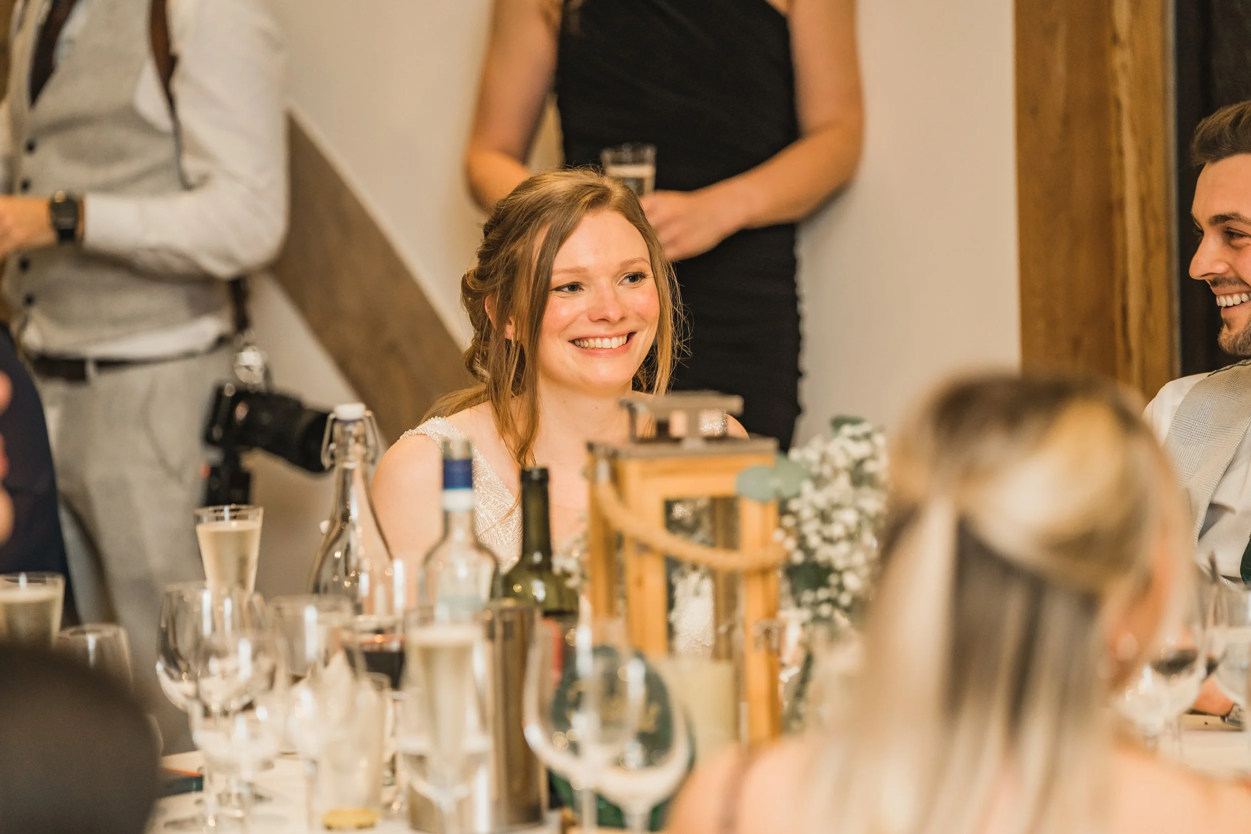 A young woman with long red hair smiling at a social gathering, seated at a table with drinks and bottles, surrounded by other people in a warmly lit room.