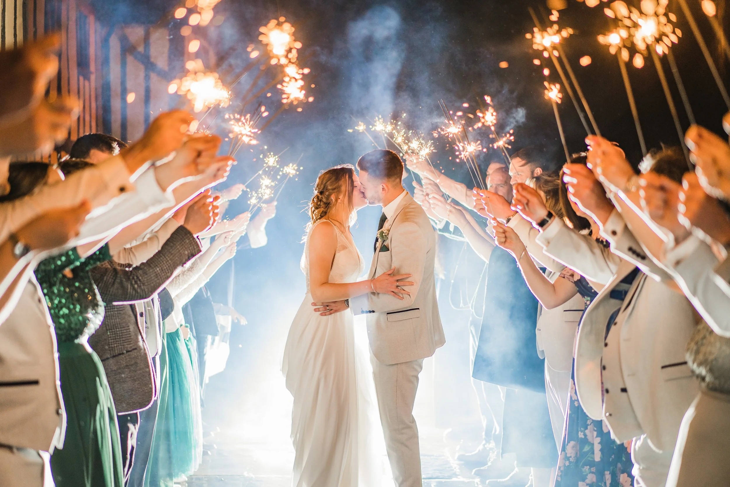 A bride and groom kissing at their wedding surrounded by guests holding sparklers at night.