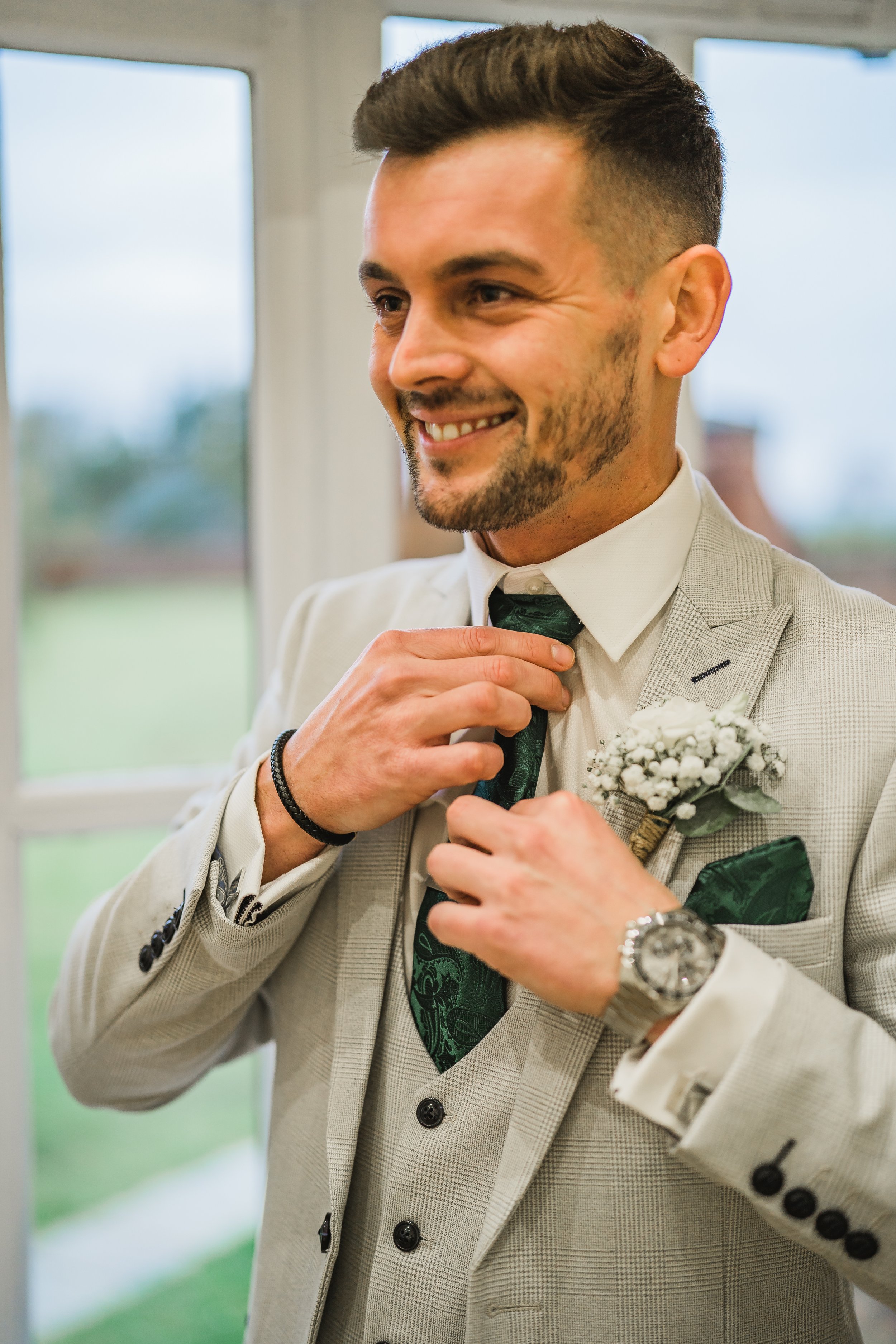 A man in a light-colored suit adjusting his tie, standing in front of windows, smiling, with a boutonniere on his lapel and a watch on his wrist.