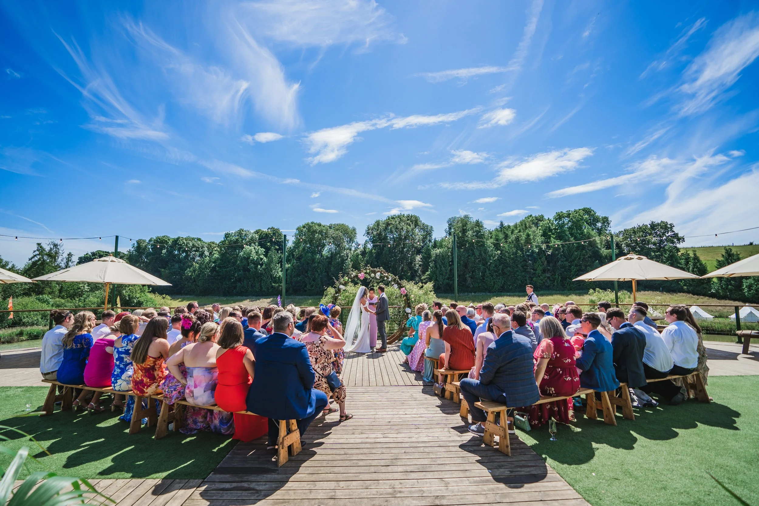 Outdoor wedding ceremony with a bride and groom exchanging vows under an arch decorated with flowers, surrounded by seated guests on wooden benches, with umbrellas, greenery, and a blue sky with clouds.