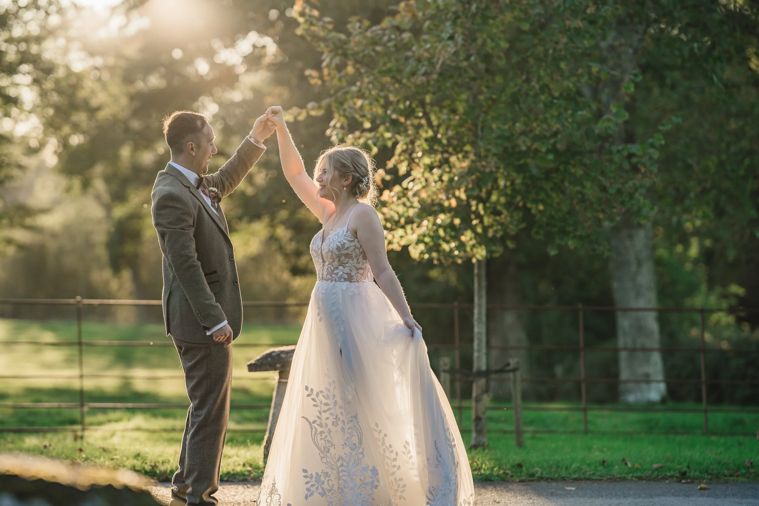 A couple is dancing outdoors during sunset, with the man in a suit and the woman in a white wedding dress, surrounded by trees.