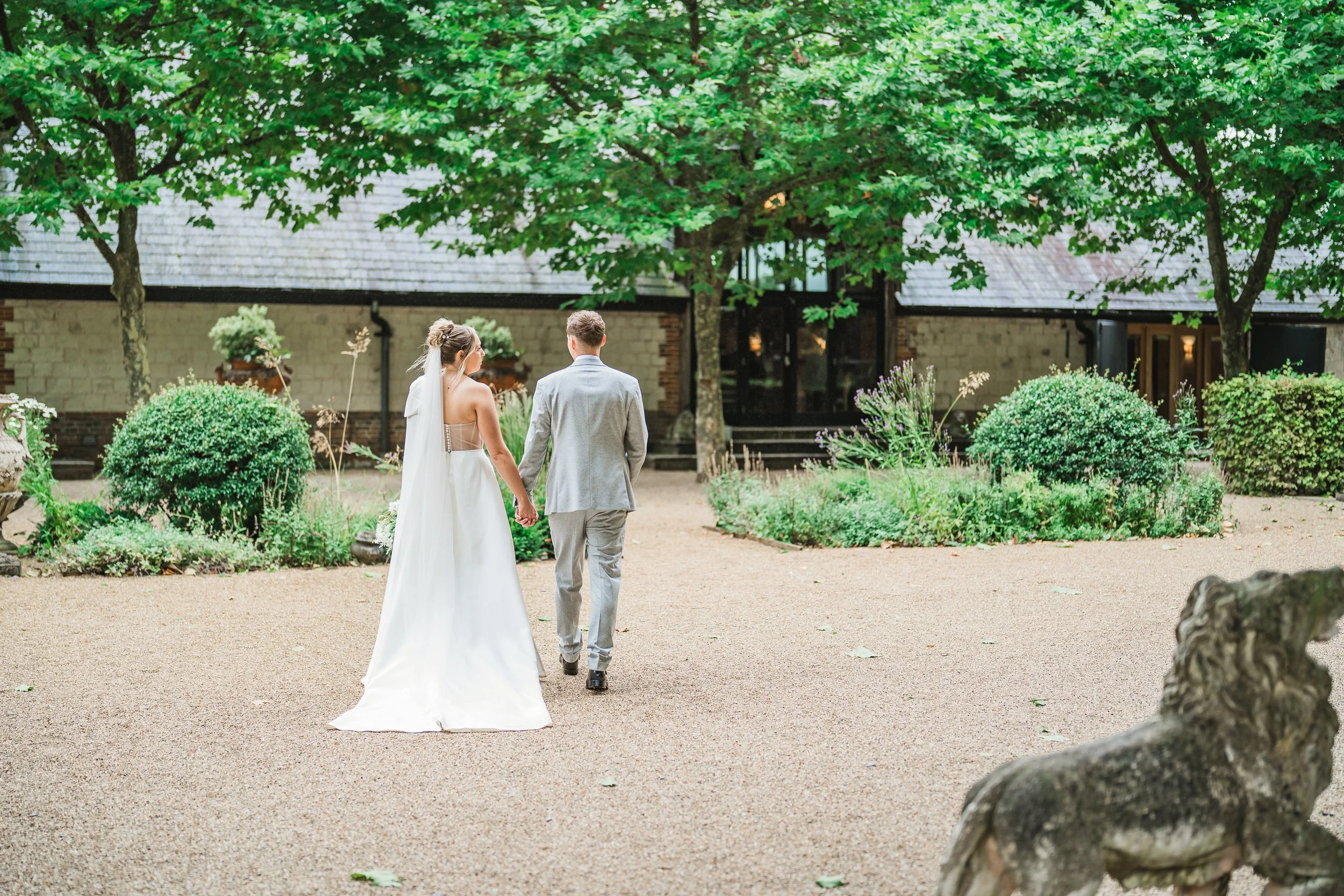 A bride and groom walking hand in hand outdoors in a garden setting, surrounded by greenery and trees.