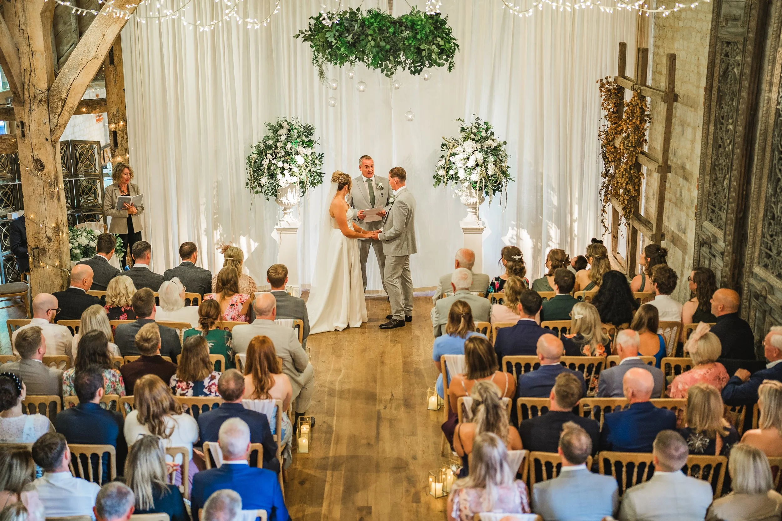 A wedding ceremony taking place indoors with a bride and groom holding hands in front of an officiant. The backdrop is decorated with white drapery, floral arrangements, and hanging glass orbs. Guests are seated facing the couple, and there are strin