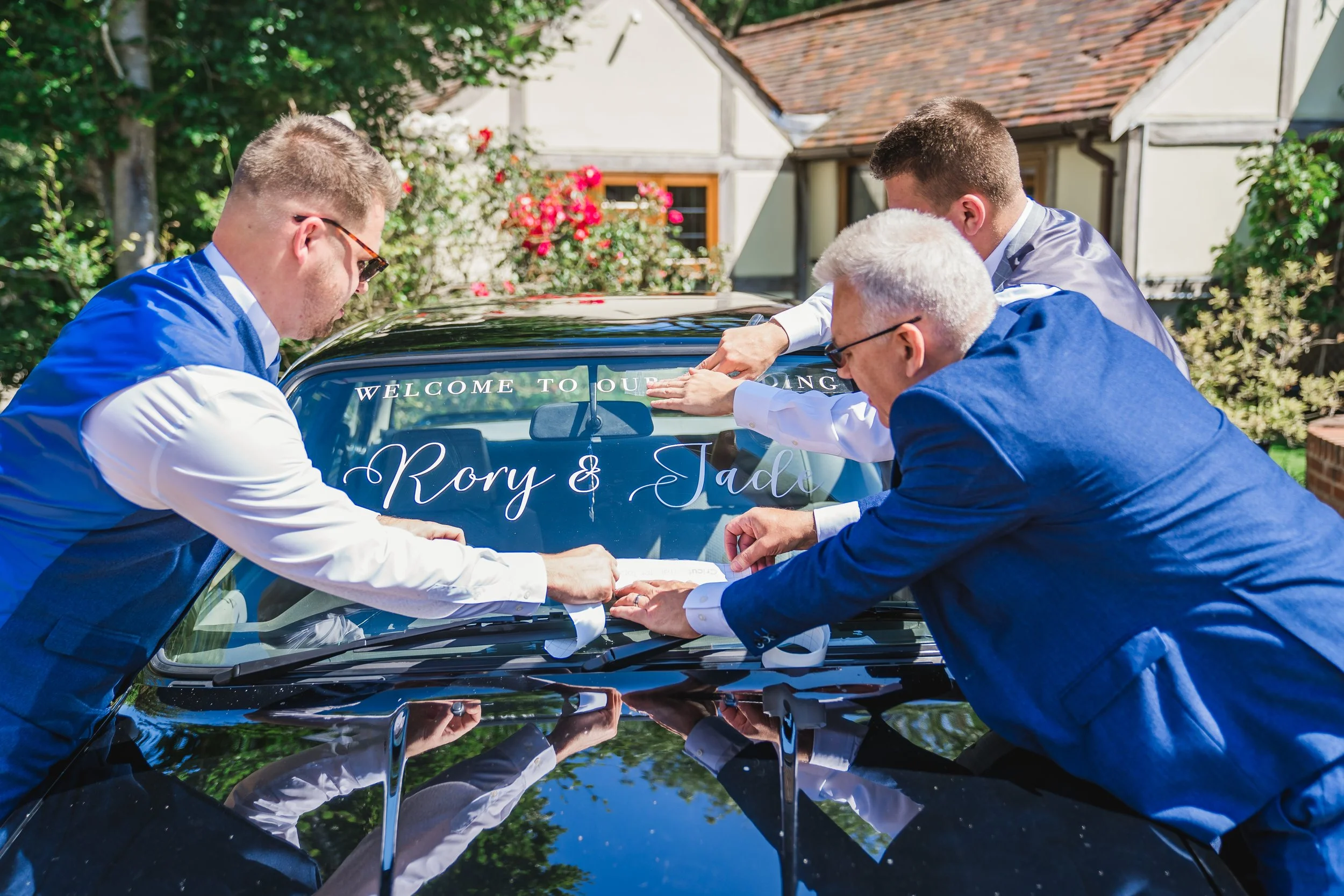 Four men in formal attire are leaning over the hood of a shiny black car decorated with a sign that says "Welcome to our wedding" and the names "Kory & Jade." They appear to be placing or adjusting a sign or decoration on the car at a wedding celebra