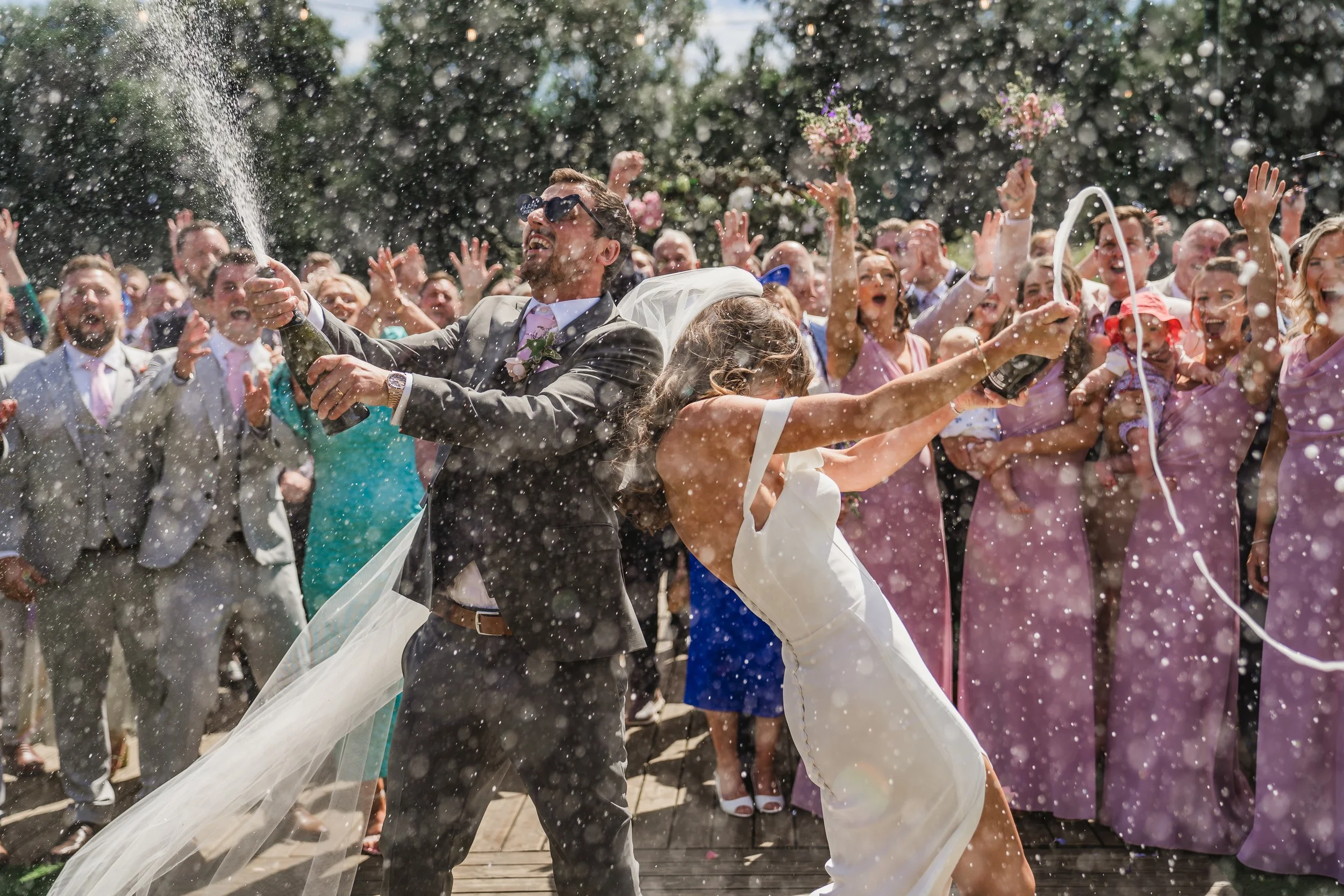 A bride and groom celebrate their wedding outdoors, spraying champagne with a large crowd of friends and family cheering around them.
