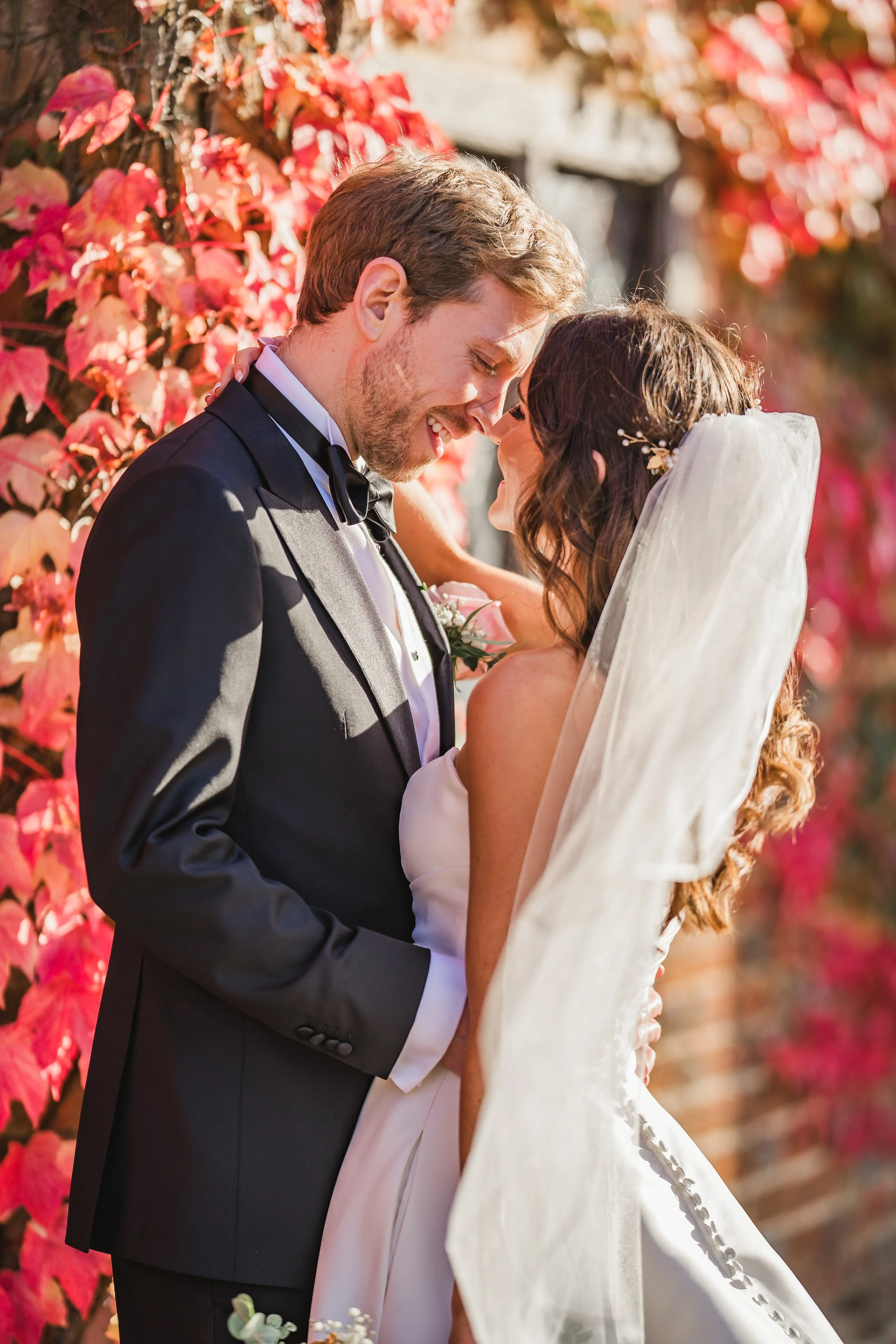 A bride and groom sharing a kiss at their wedding outdoors, with pink and red foliage in the background.