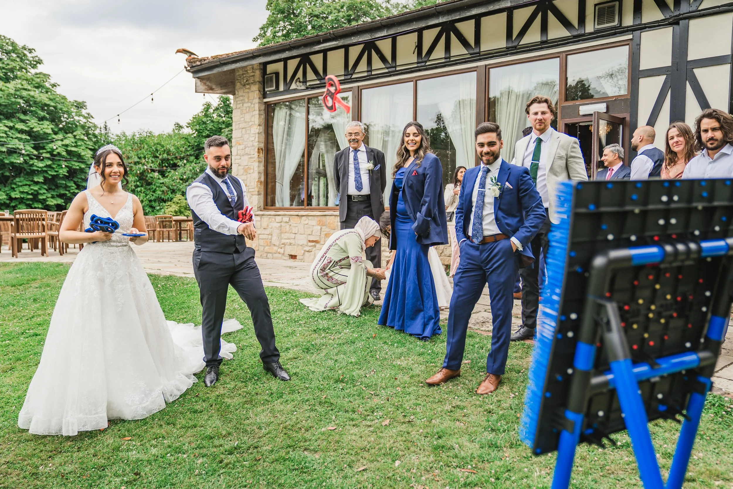 A bride in a white wedding dress holding a plate and a groom in a blue vest and tie next to her. Several people in formal attire stand outside a building with curtains, preparing for a wedding ceremony as a camera is set up to film the event.