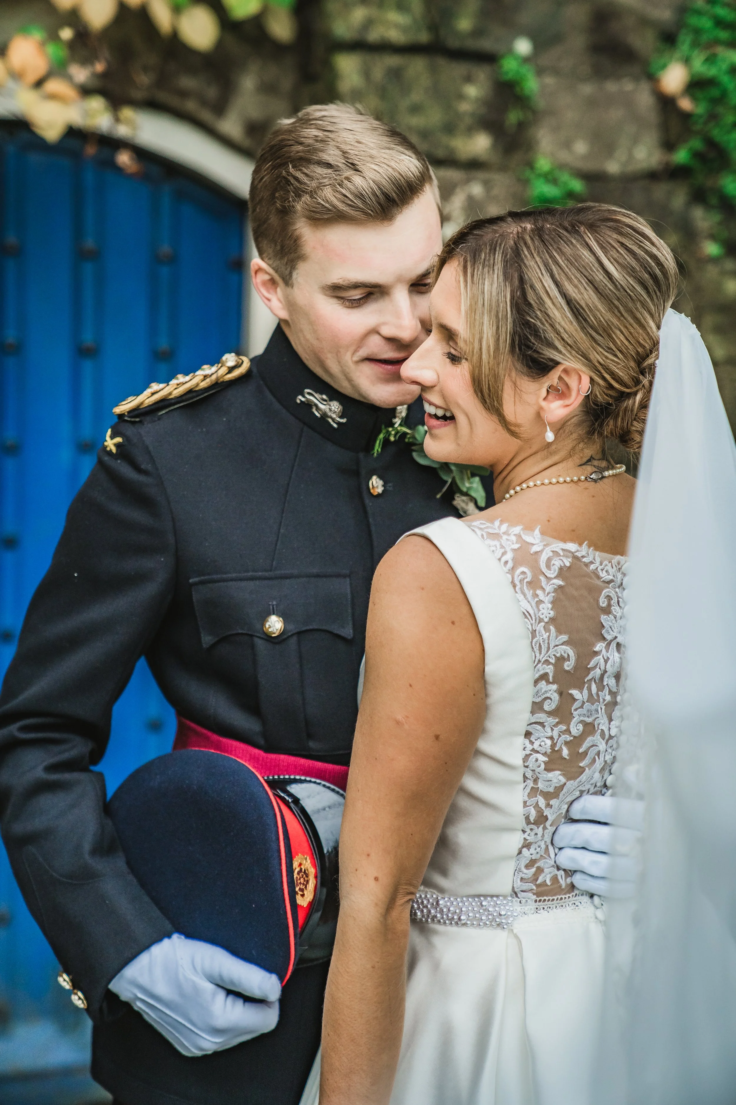 A couple, dressed in wedding attire, sharing a kiss at their wedding, with the groom in a military uniform and the bride in a lace wedding dress with pearl jewelry.