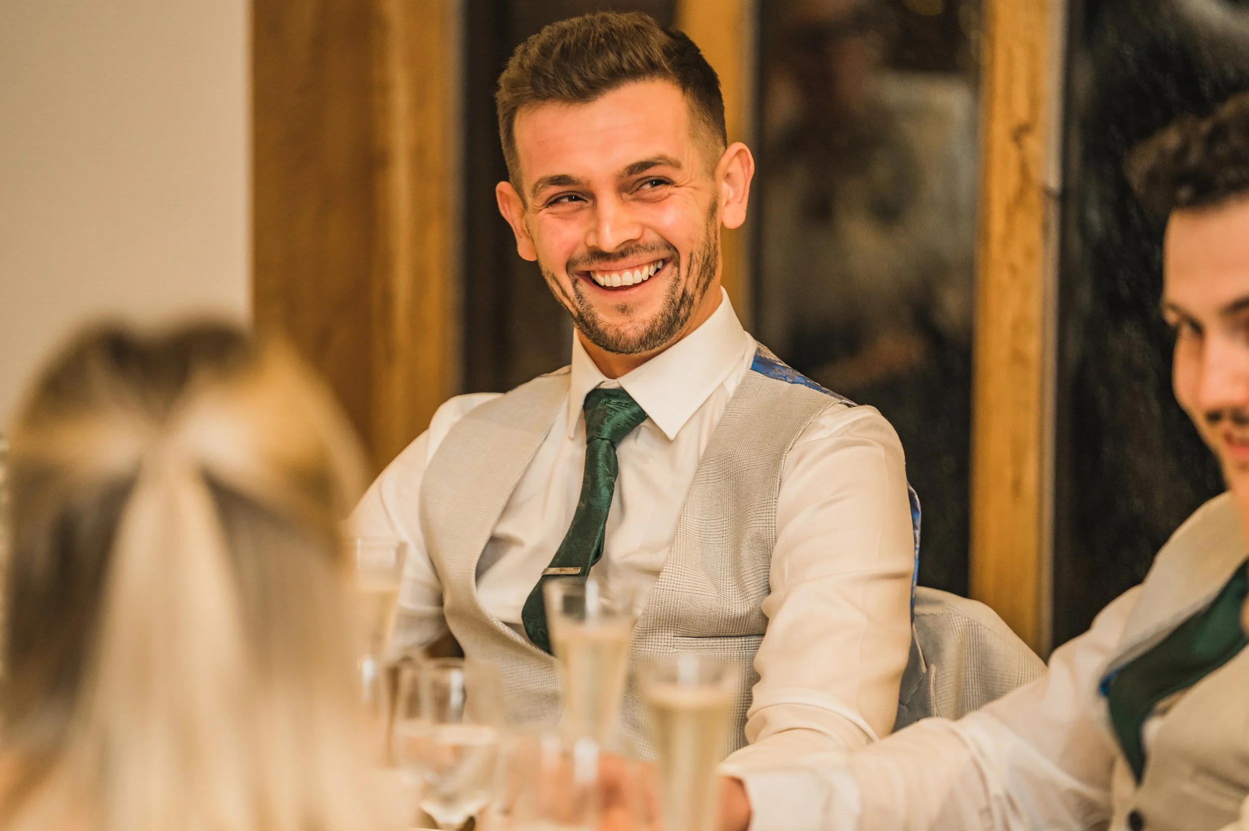 A man with short dark hair and a beard, dressed in a white shirt and light-colored vest, smiling at a gathering with other people, with glasses of wine and a candle on the table.