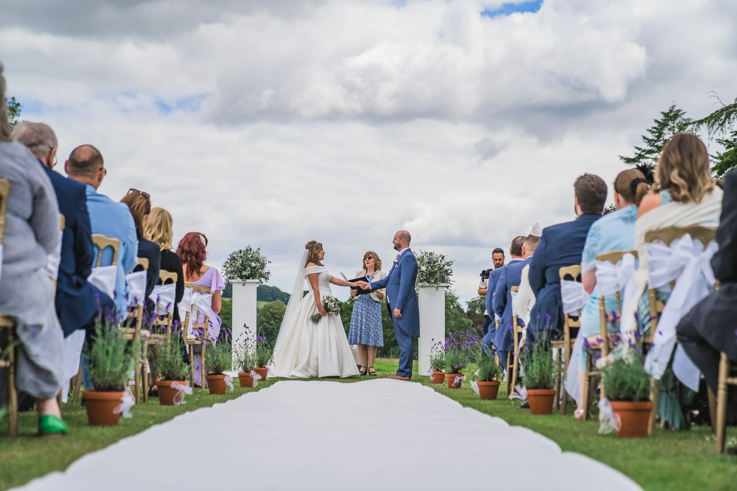 An outdoor wedding ceremony with a bride and groom holding hands in front of officiant, surrounded by seated guests on either side of an aisle decorated with potted plants and lavender flowers, under a cloudy sky.