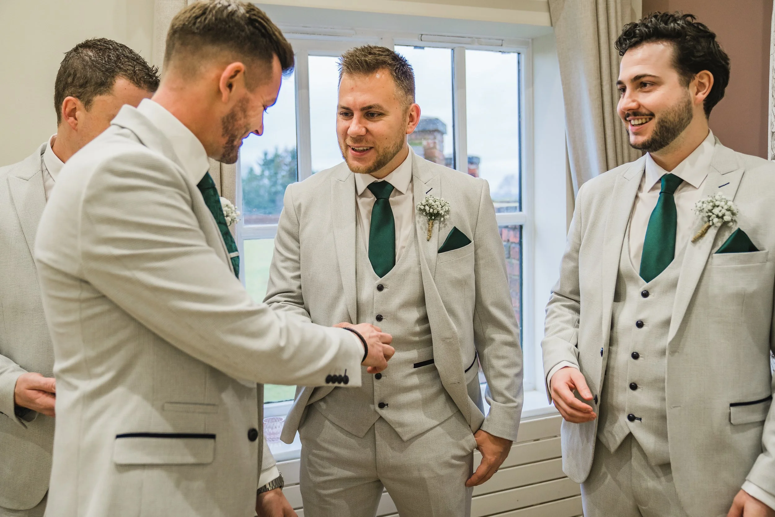 A group of five men dressed in light-colored suits with dark green ties and boutonnières are engaged in a handshake or greeting in a room with a window and beige curtains.
