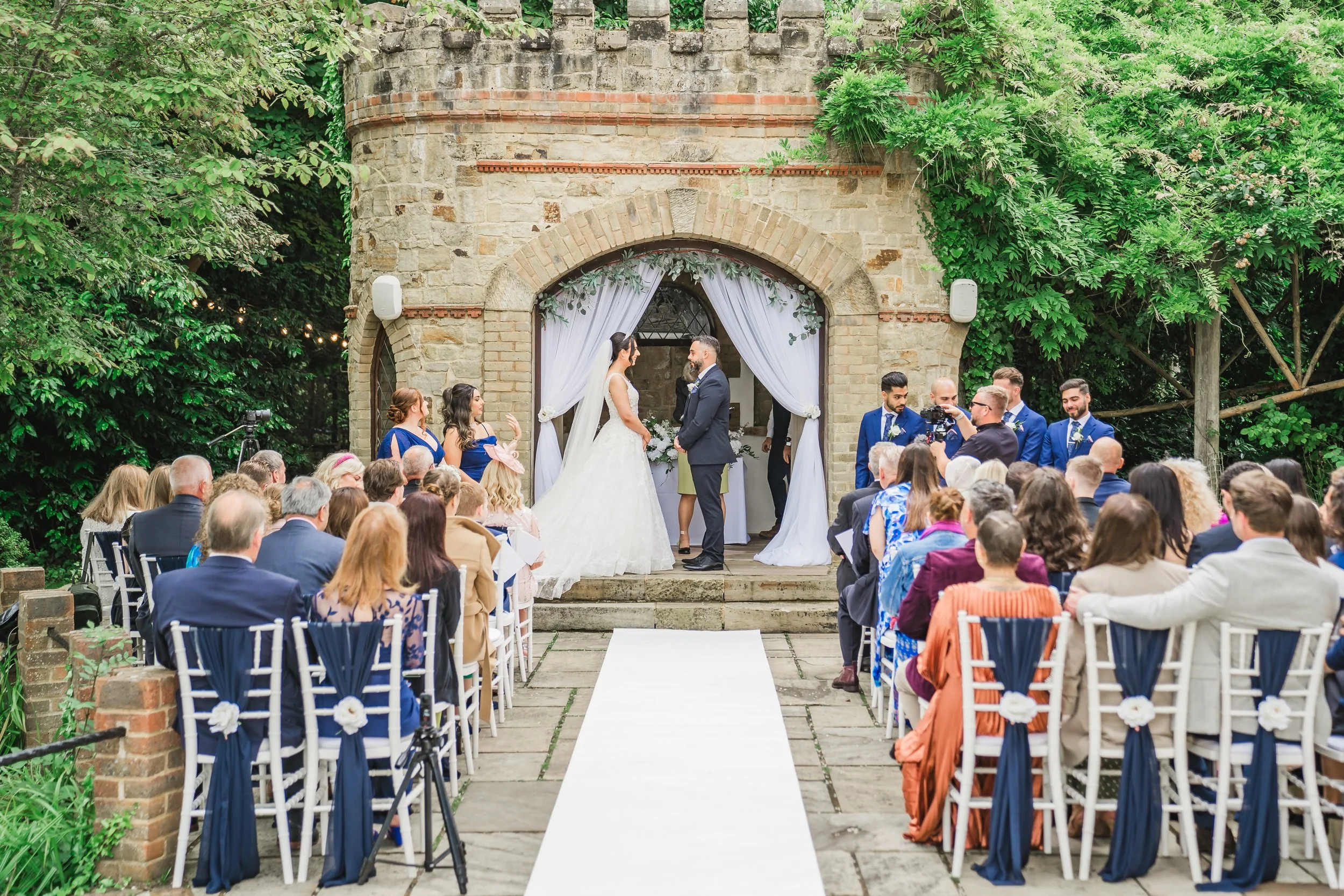 A wedding ceremony outdoors with the bride and groom standing under a decorated arch, surrounded by guests seated on white chairs, near a stone building with greenery.