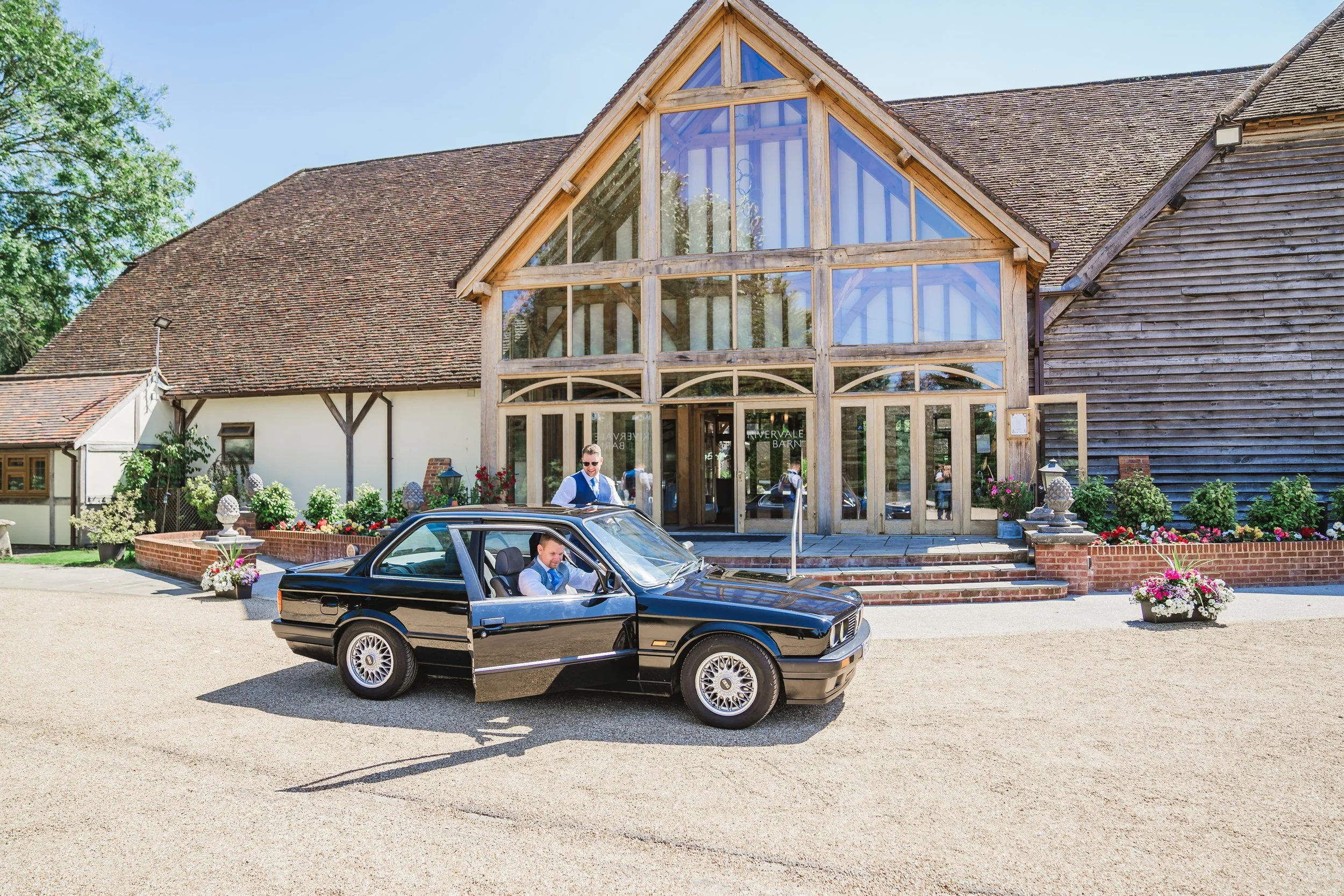 A black vintage car with two men dressed in formal white shirts and blue vests, one driving and the other standing next to it, parked in front of a rustic barn-style building with large glass windows and flowers outside Rivervale Barn