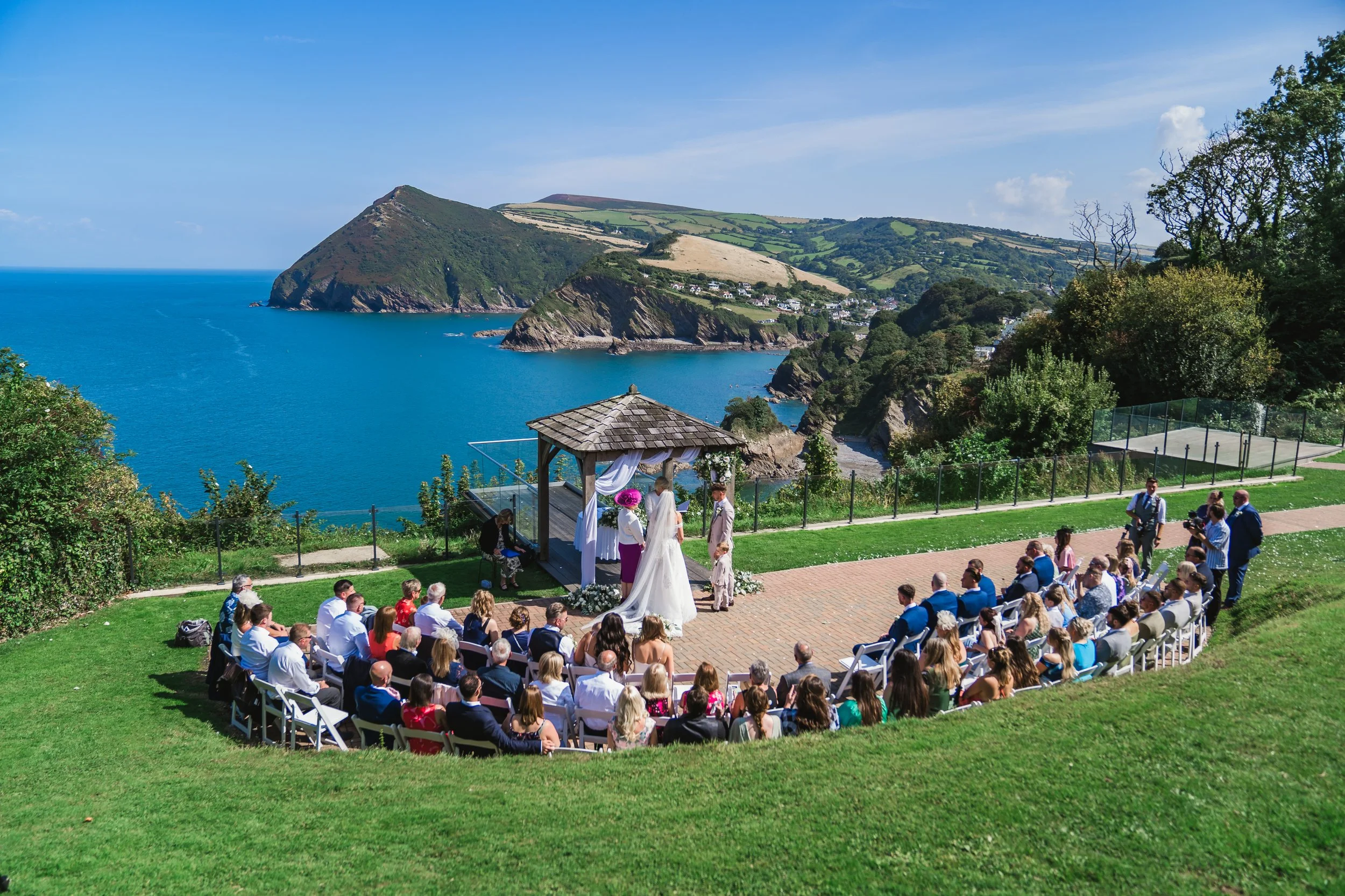 Outdoor wedding ceremony near a coastline with green hills and water, guests seated in chairs, officiant and couple at altar with white drapery and flowers, clear sky.