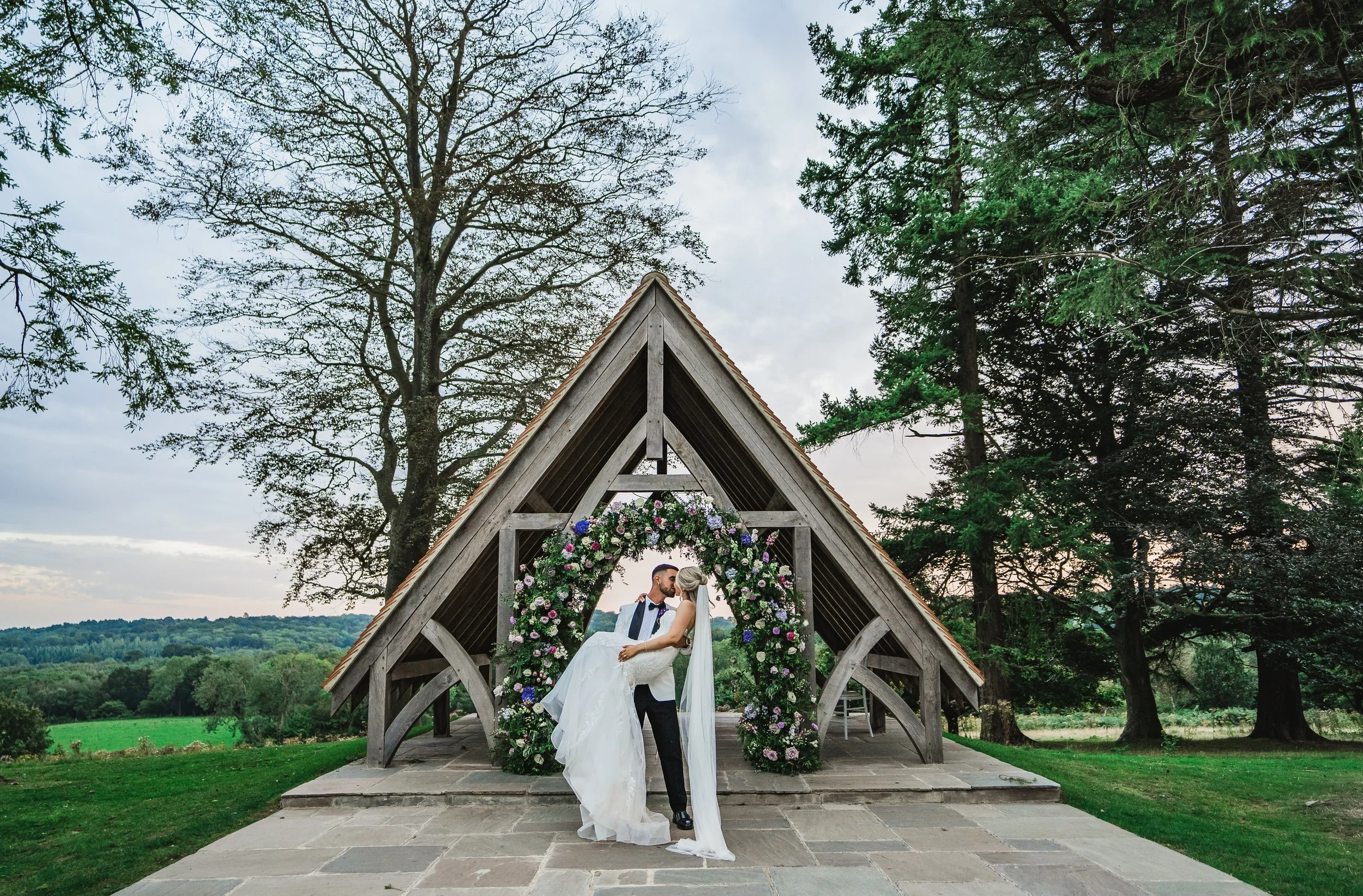 A bride and groom sharing a kiss under a floral wedding arch at an outdoor wedding ceremony.